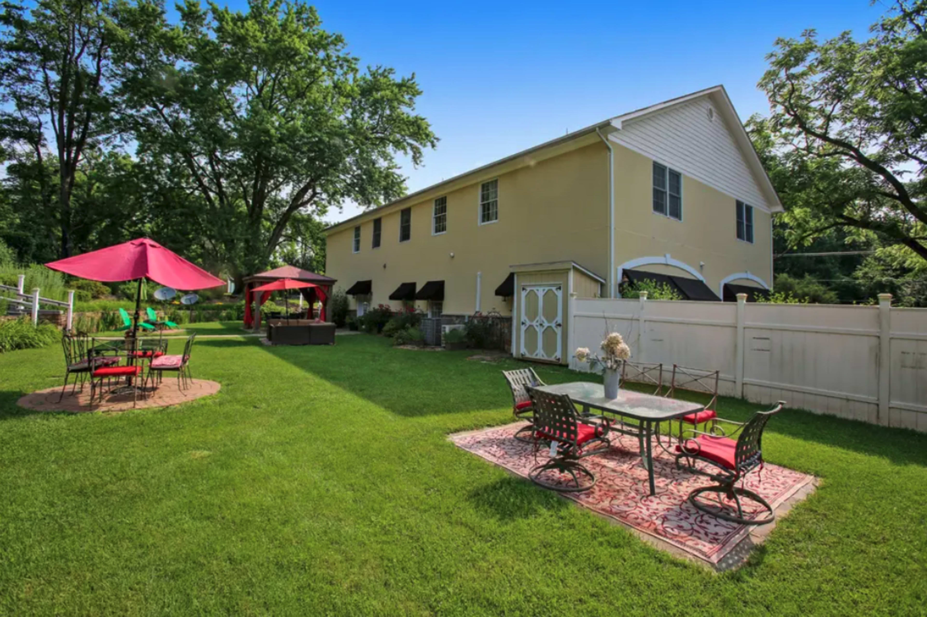 A spacious backyard with a lawn, patio furniture, umbrellas, and a shed behind a two-story yellow house, surrounded by trees.