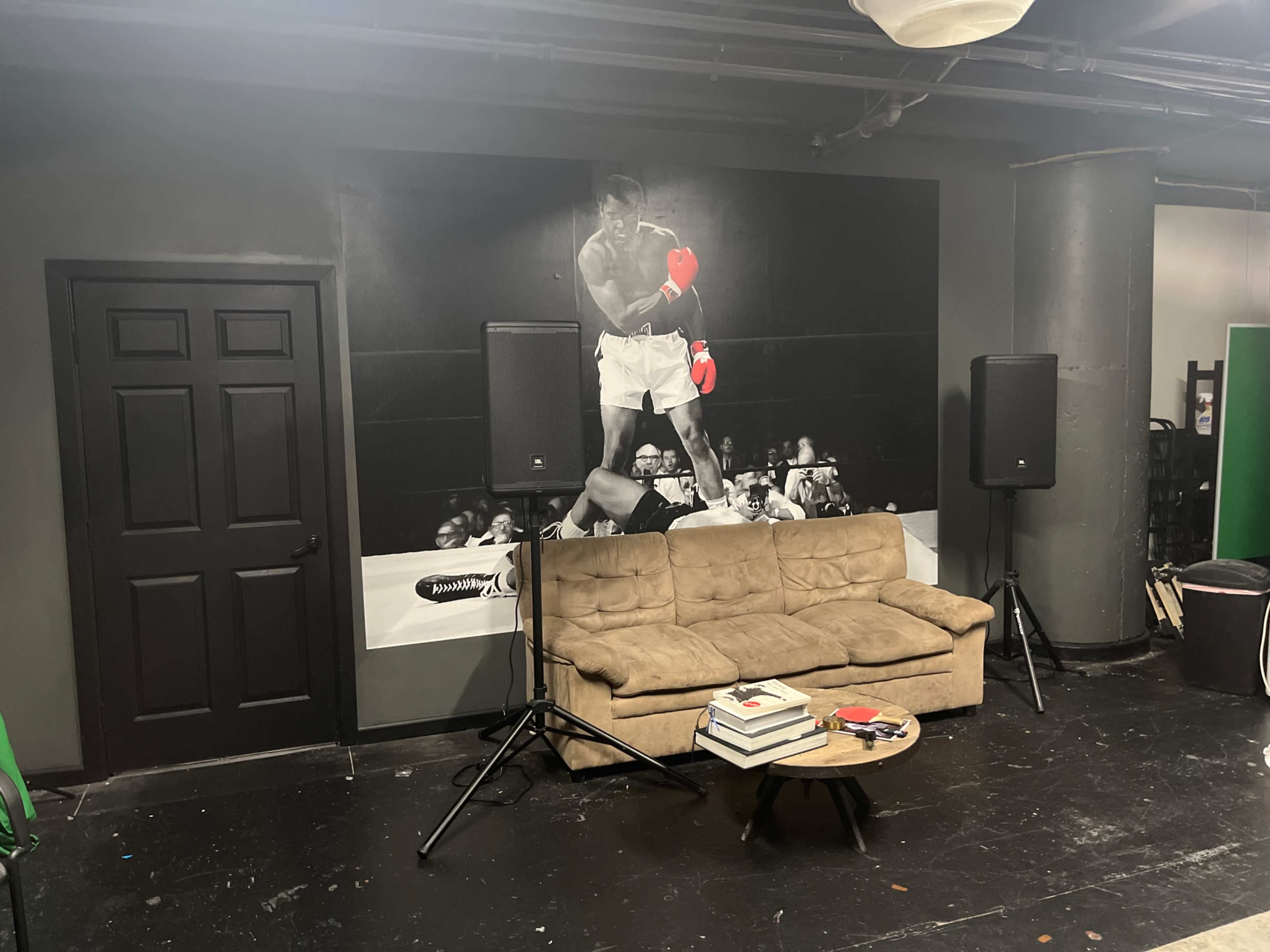 A room with a large mural of a boxer on the wall, a beige couch, a small table with books, and two speakers positioned beside the couch.