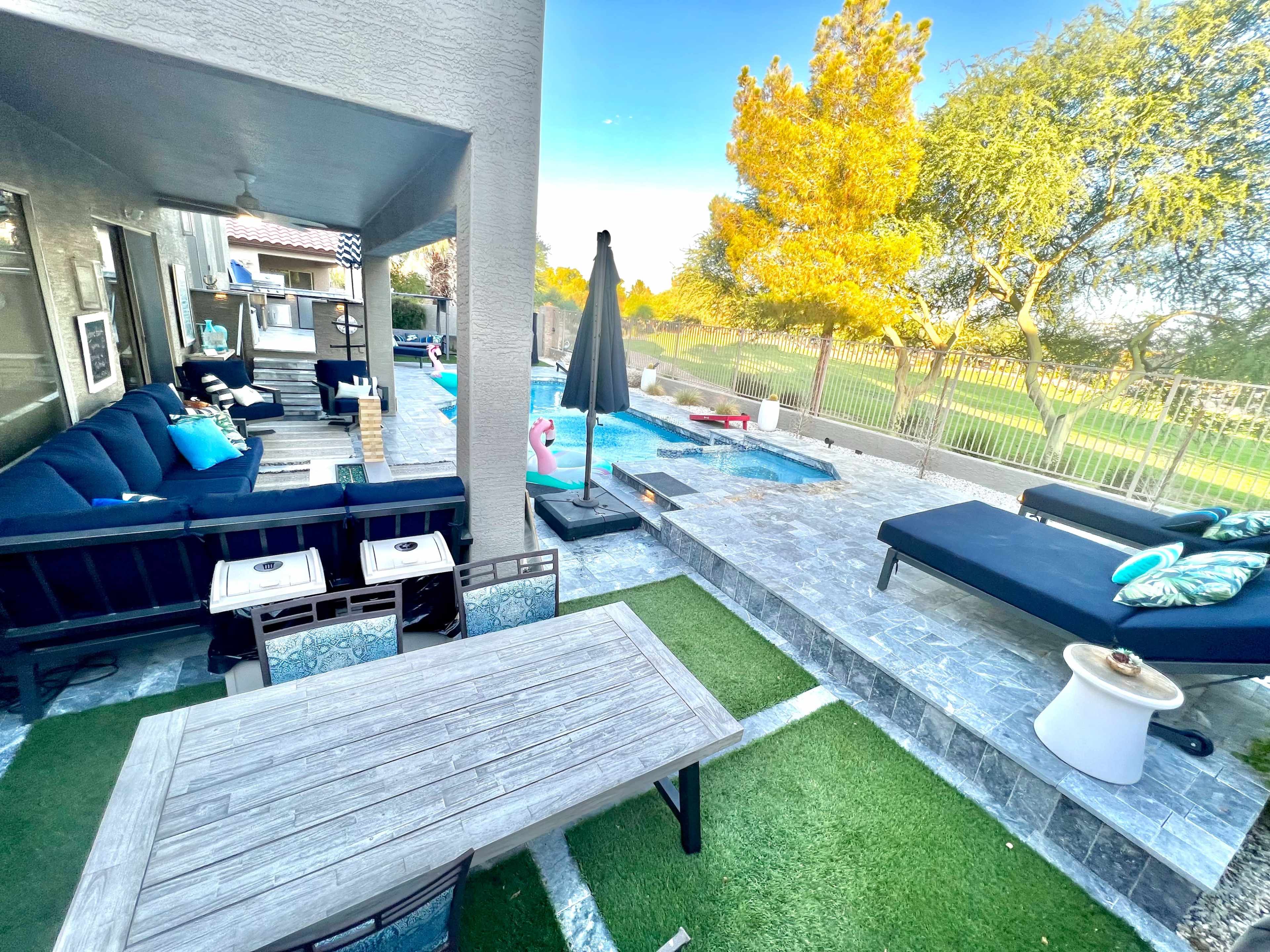 The image shows a backyard patio with a pool, a seating area with a large sofa, a dining table, and a sun lounger surrounded by greenery.