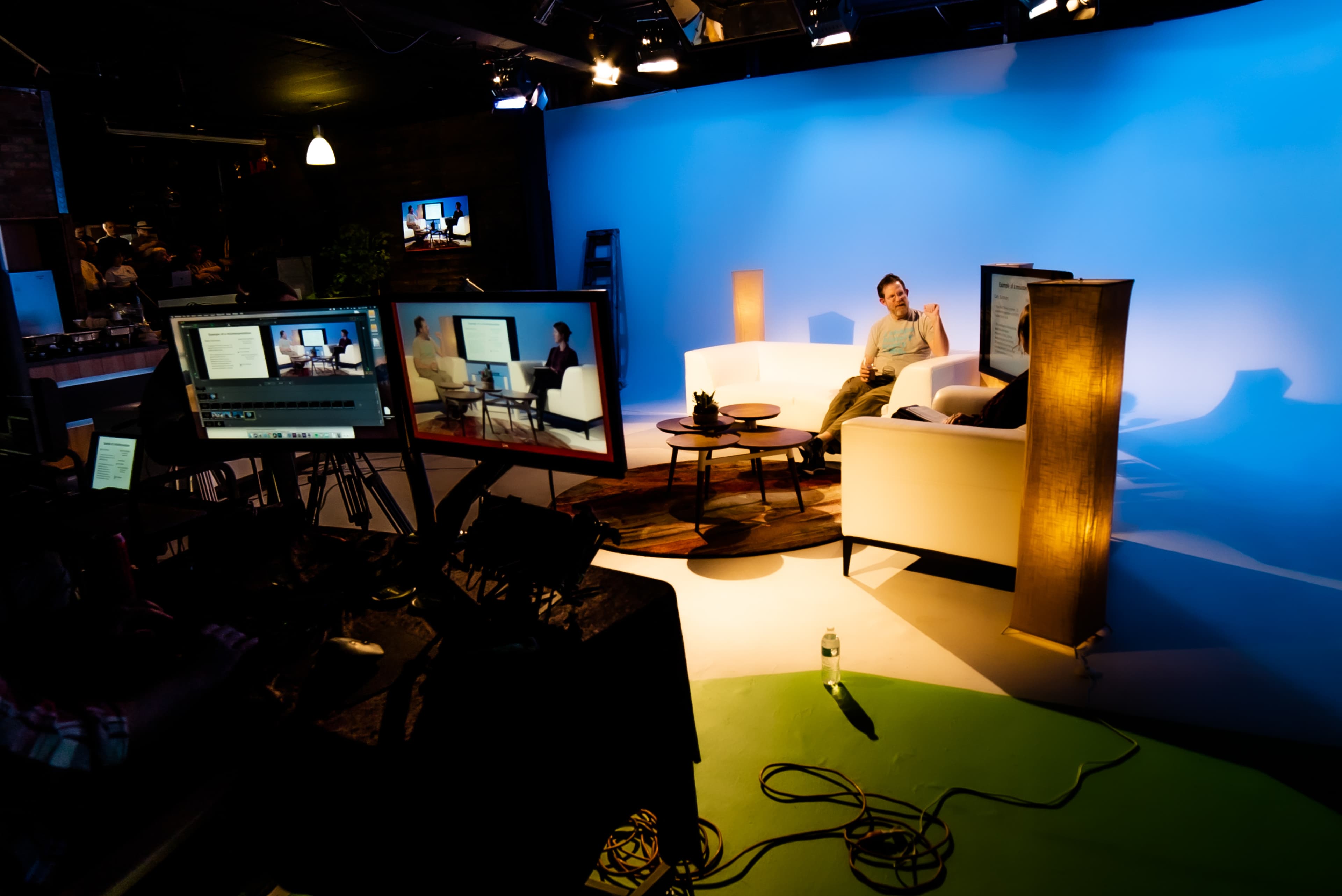 A person sits on a white sofa in a television studio setup with multiple screens and lights against a blue backdrop.