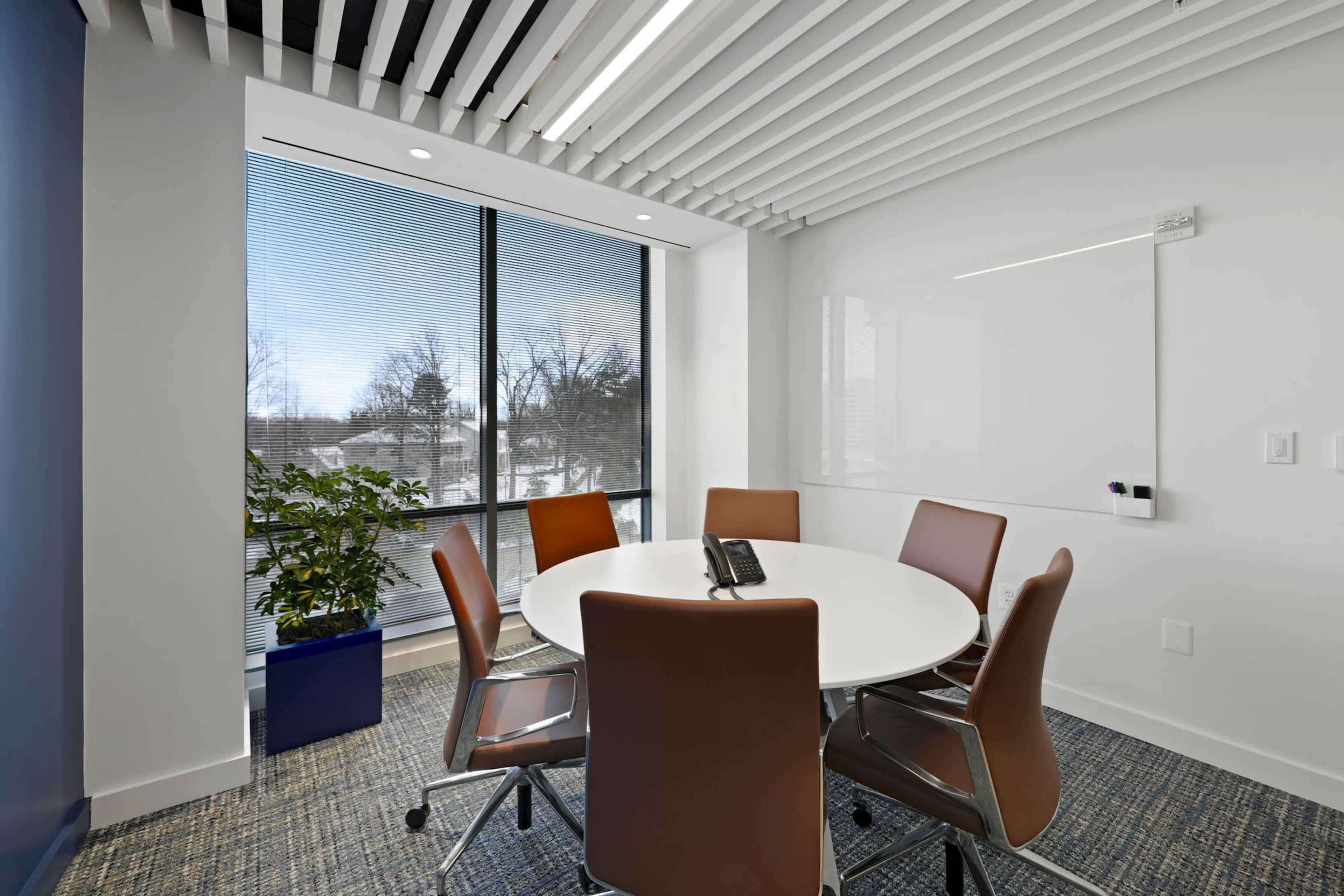 A modern conference room features a circular table surrounded by brown chairs, large windows with blinds, and a potted plant in a blue planter.