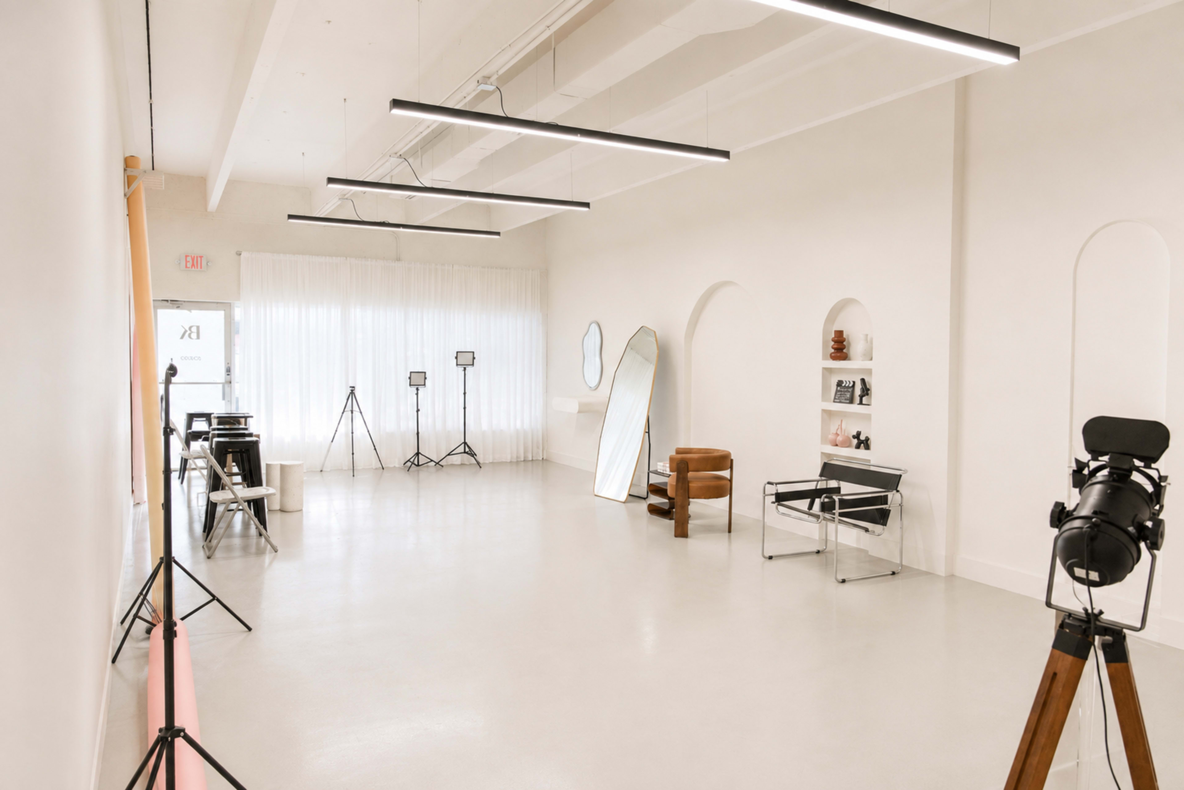 The image shows a bright, modern studio space with minimalist furnishings, including a mirror, a chair, and equipment stands arranged along the walls.