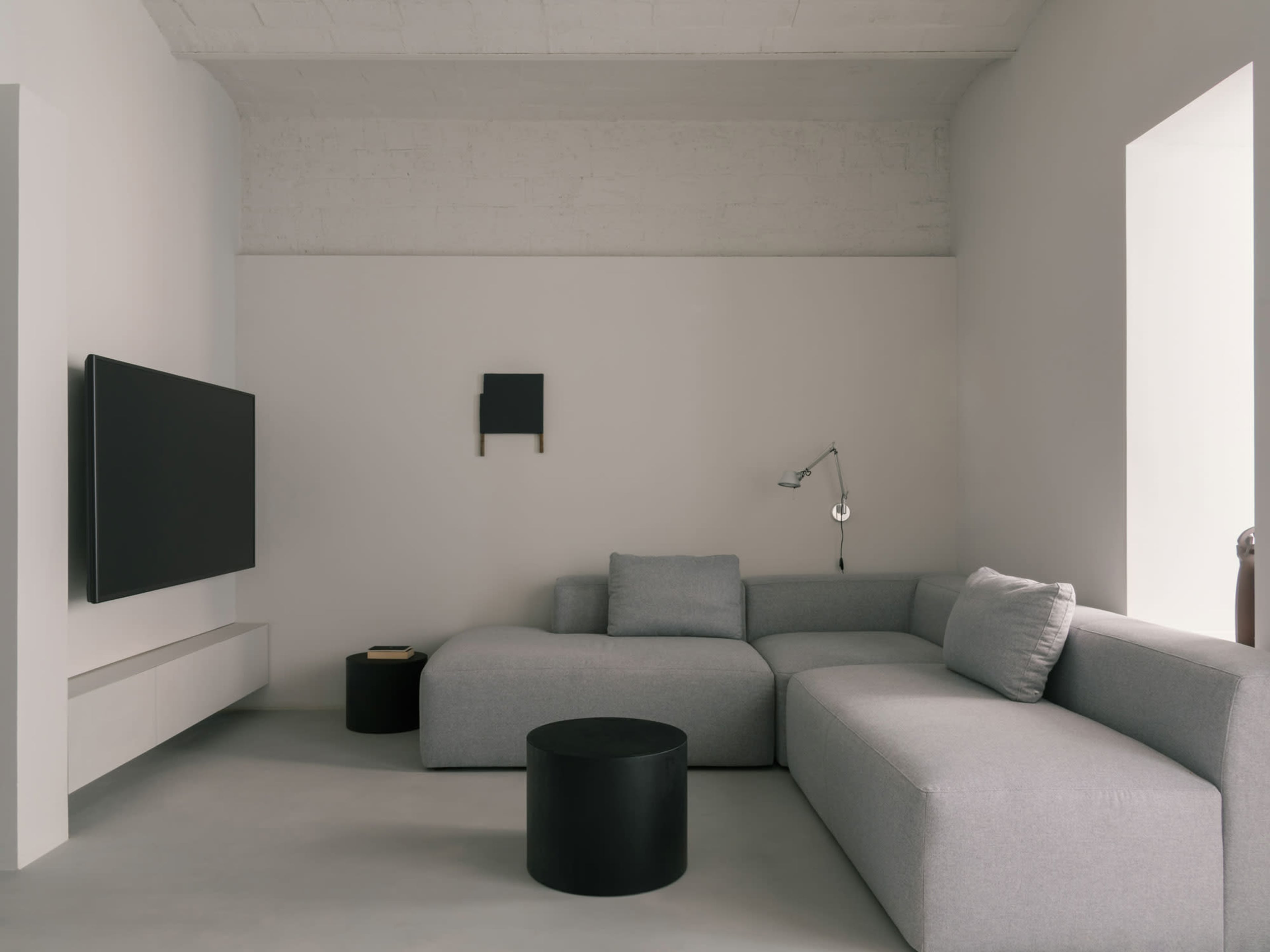 The image shows a minimalist living room with a gray sectional sofa, a black round coffee table, and a wall-mounted television.