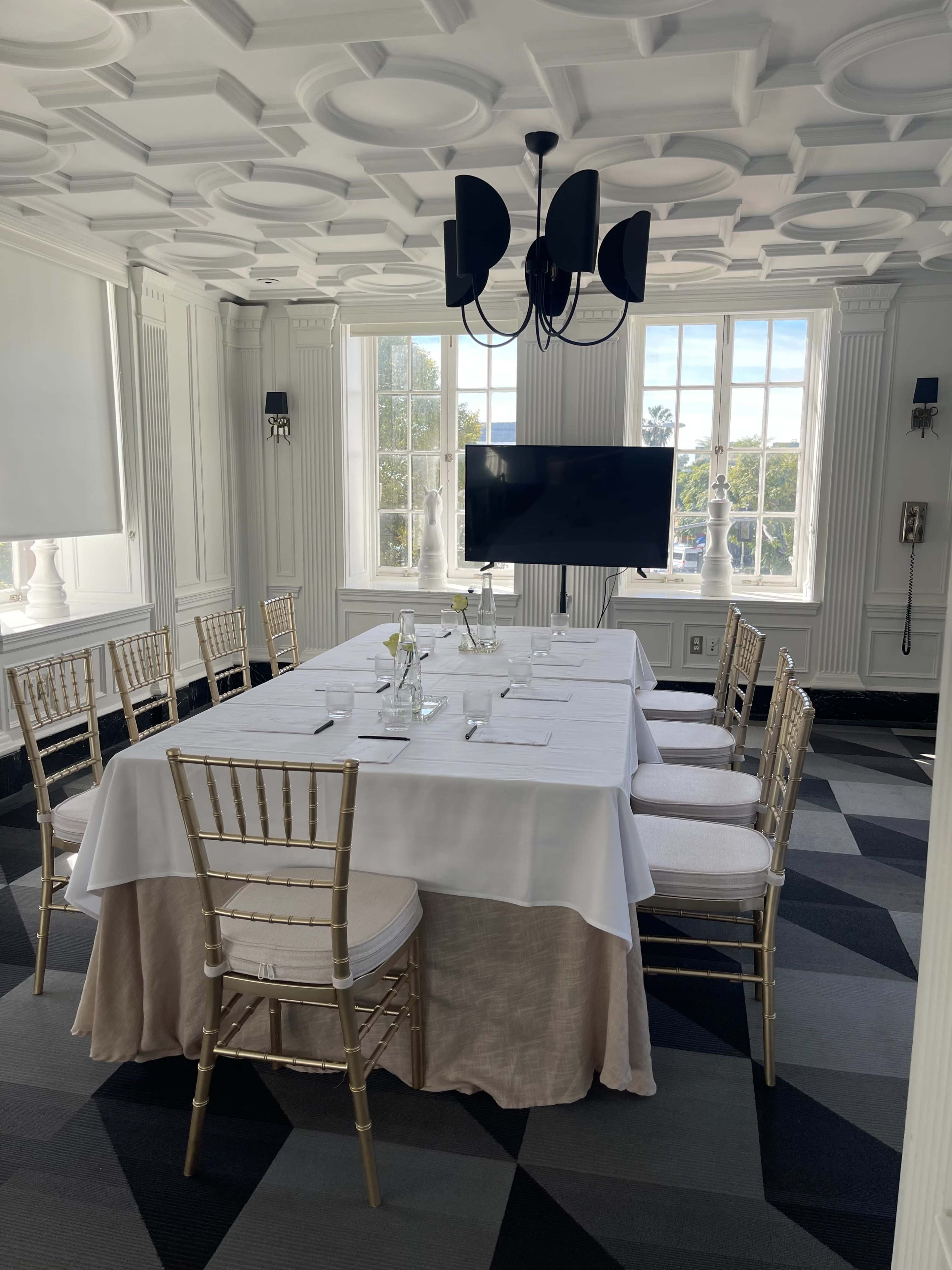 The image shows a well-lit meeting room with a large table set for a formal gathering, surrounded by gold-colored chairs and featuring a screen at one end.
