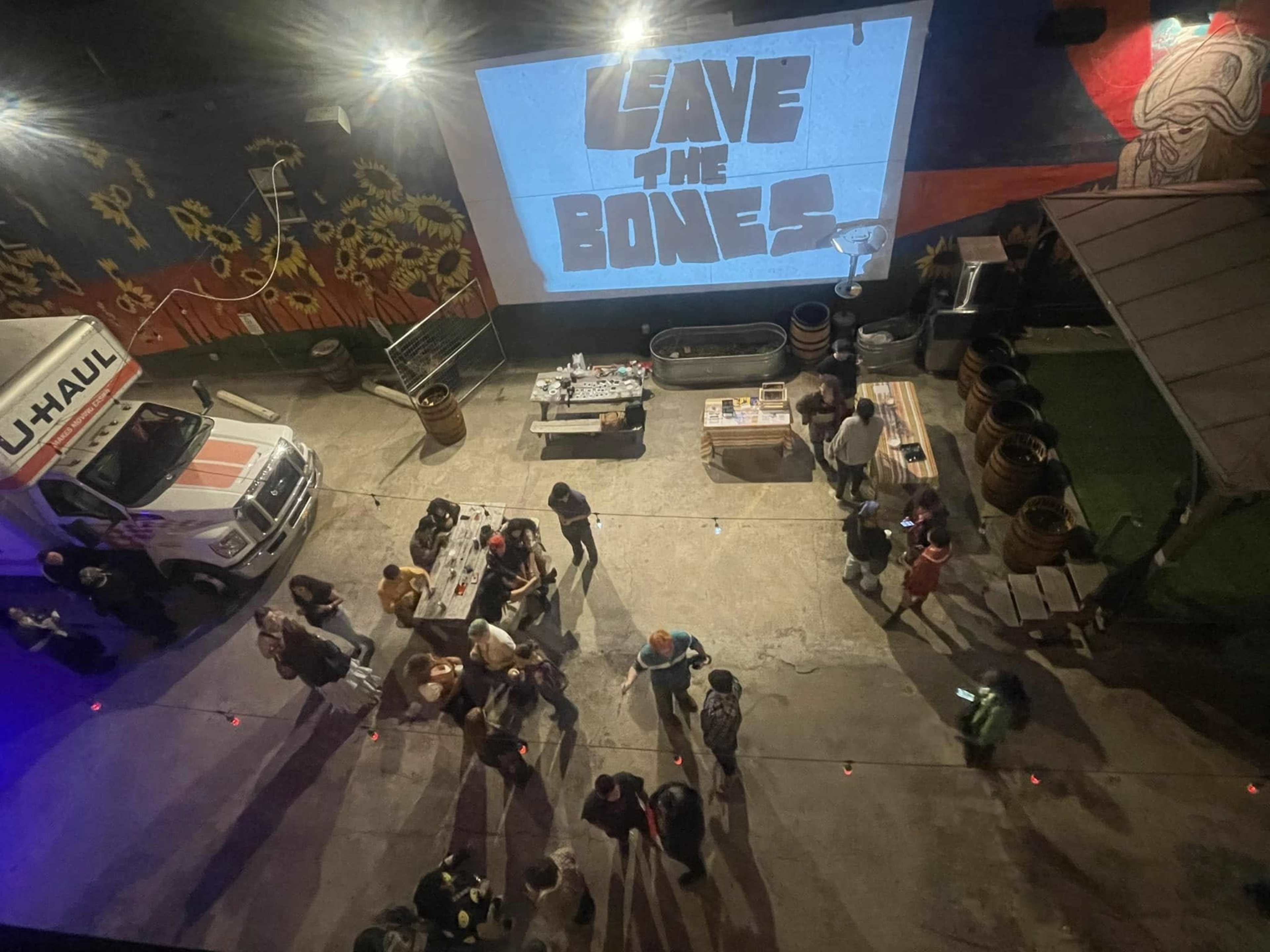 The image shows a lively outdoor gathering with people mingling around tables, a U-Haul truck parked nearby, and a large screen displaying "SAVE THE BONES."