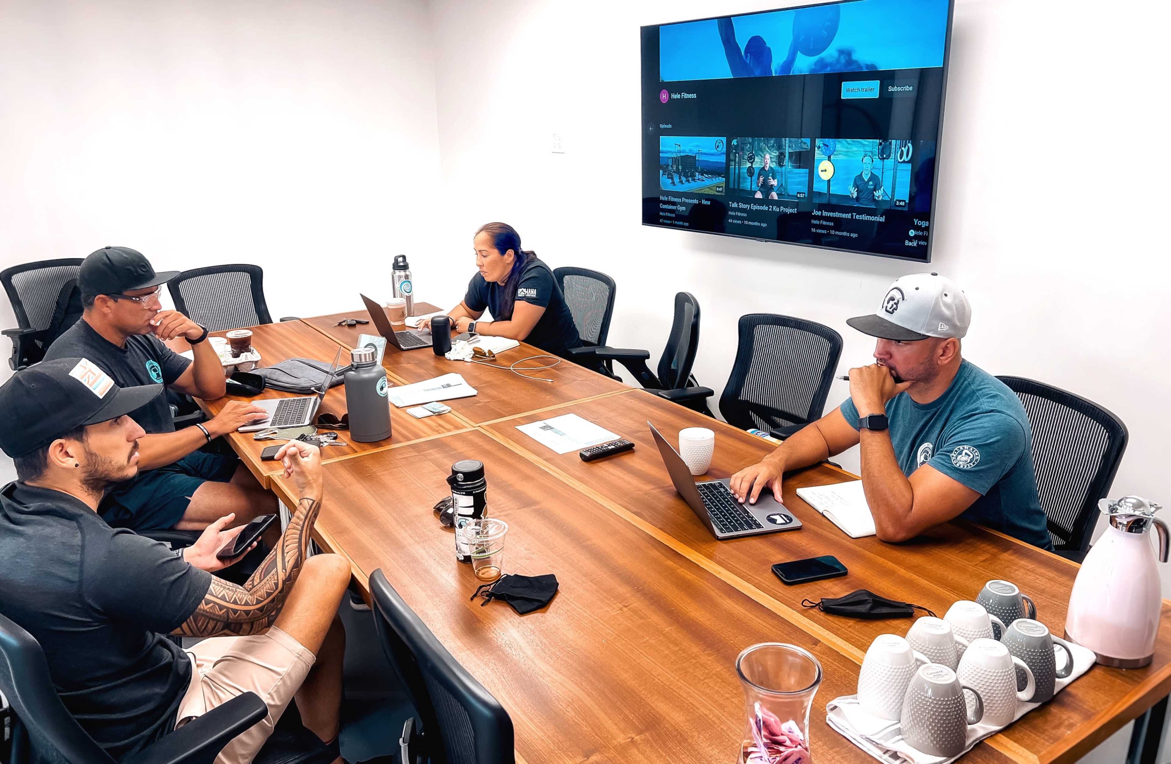 Four people are seated around a large wooden conference table in a meeting room, with a screen displaying content mounted on the wall.