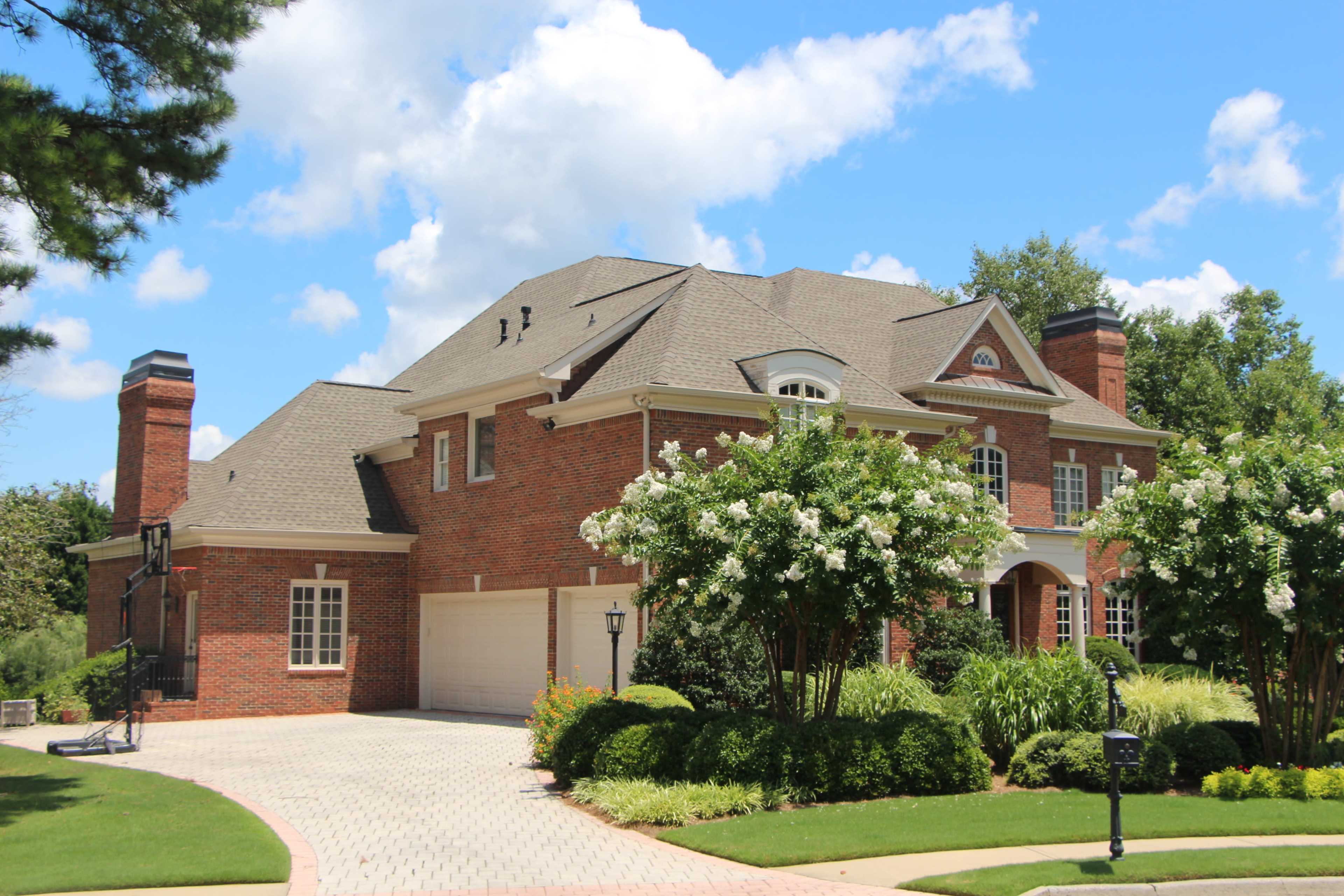 The image shows a large brick house with a multi-gabled roof, surrounded by well-maintained landscaping and a paved driveway.