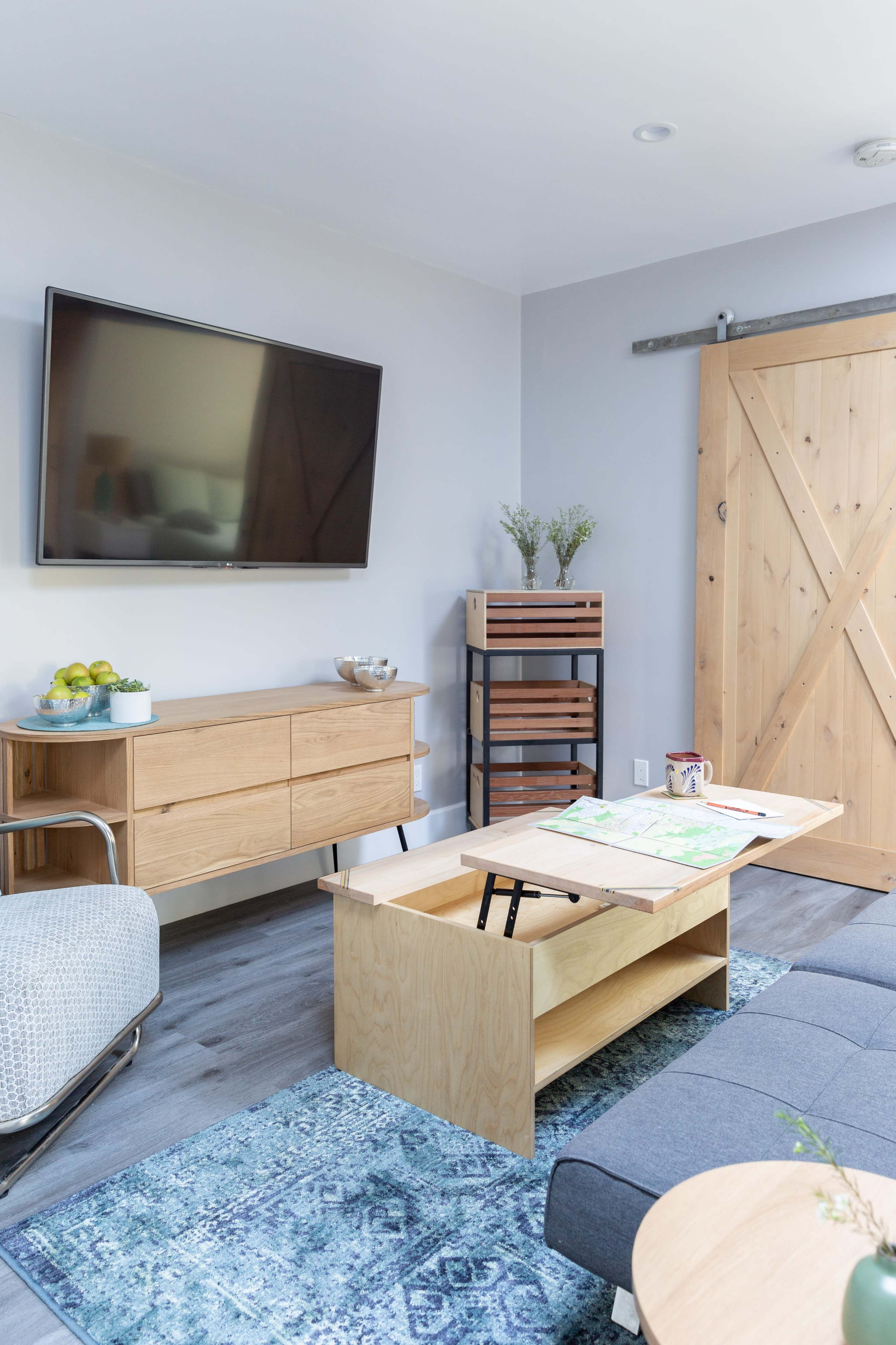 The living room features a modern television mounted on the wall above a wooden sideboard, with a sliding barn door and a coffee table that can be converted for different uses.