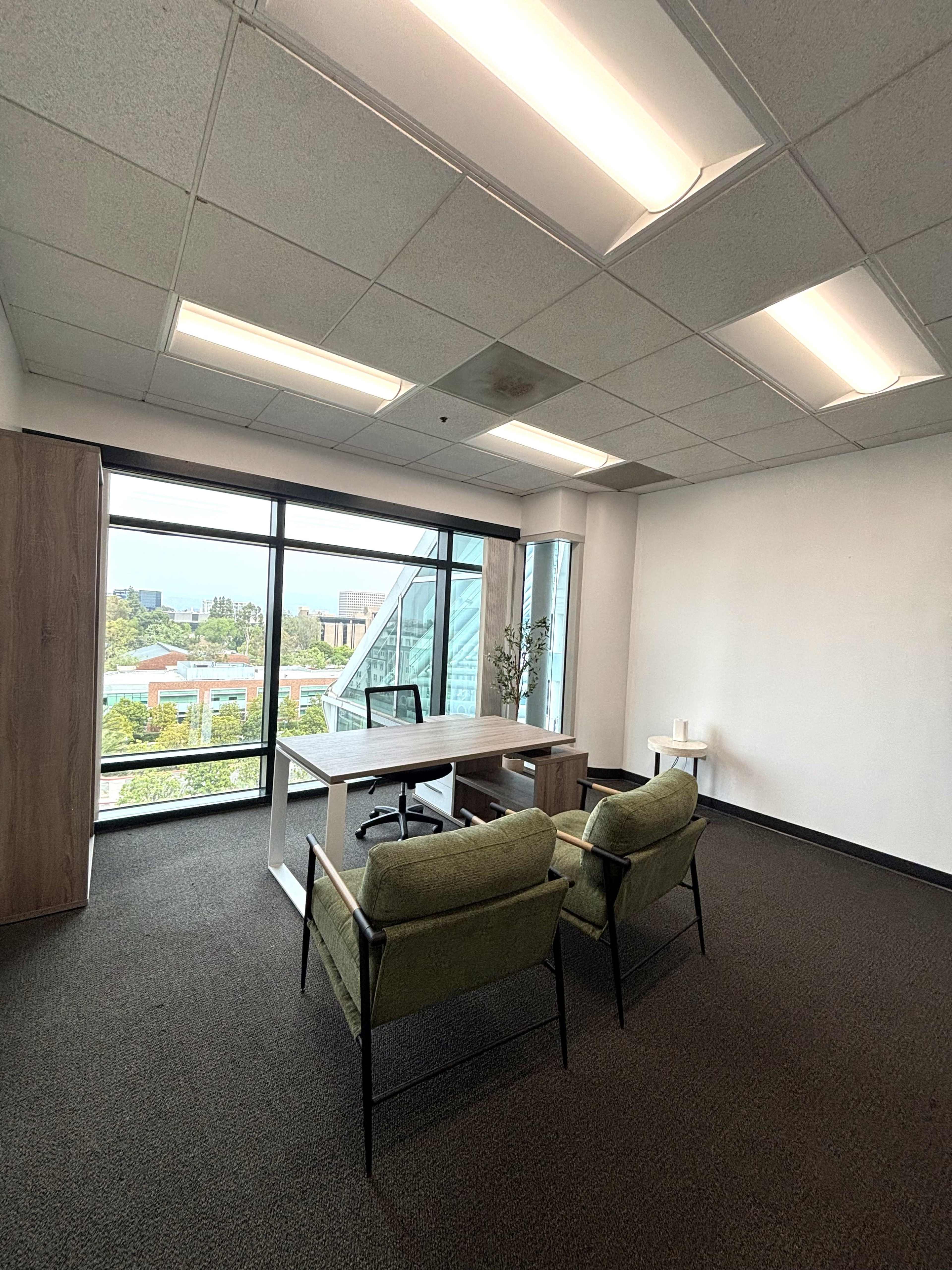 The image shows a modern office space with a large window, a desk, two green chairs, and minimal decorations.
