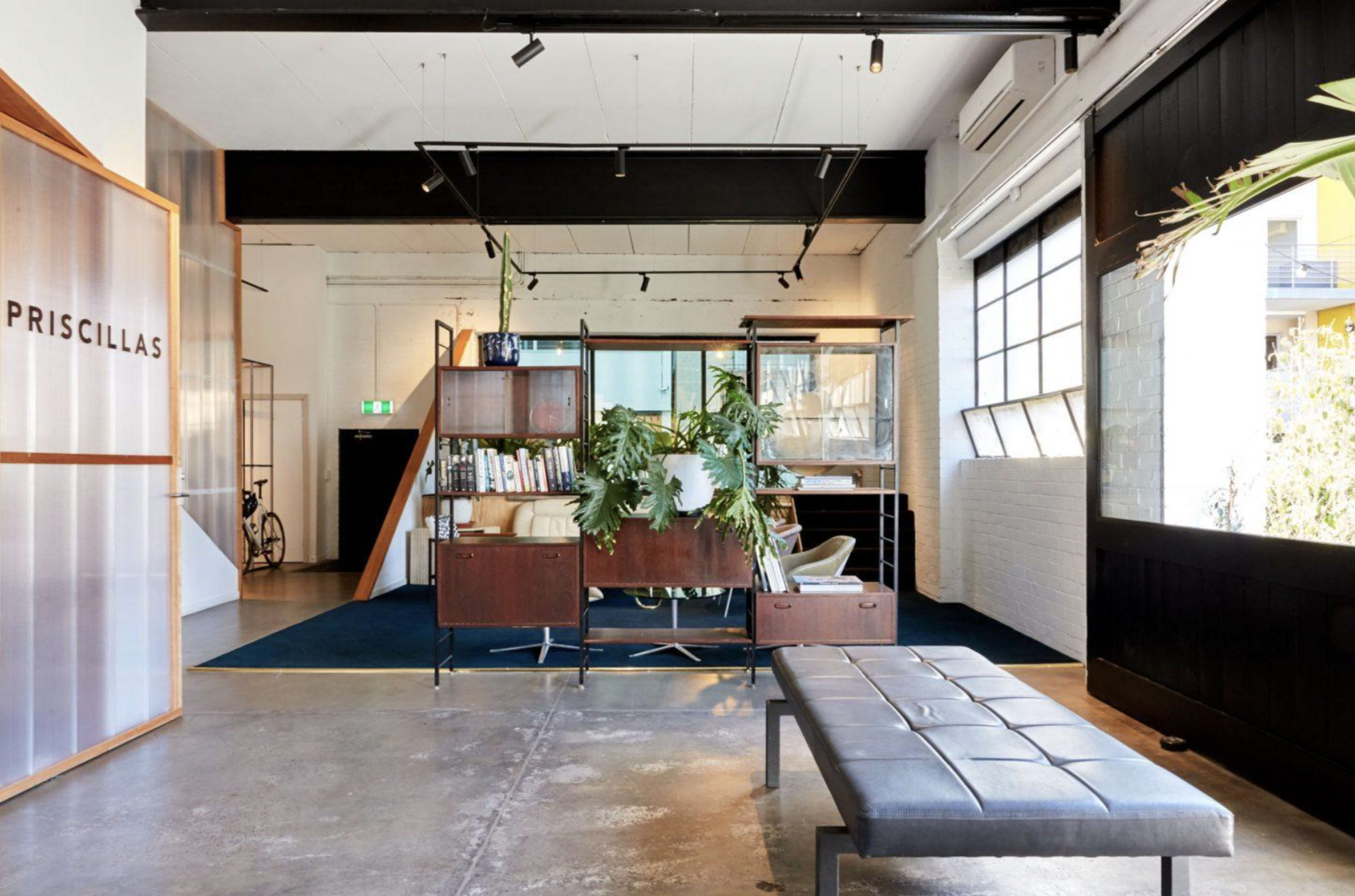 A modern office space with industrial design elements, featuring a seating area, a bookshelf, and large windows allowing natural light to enter.