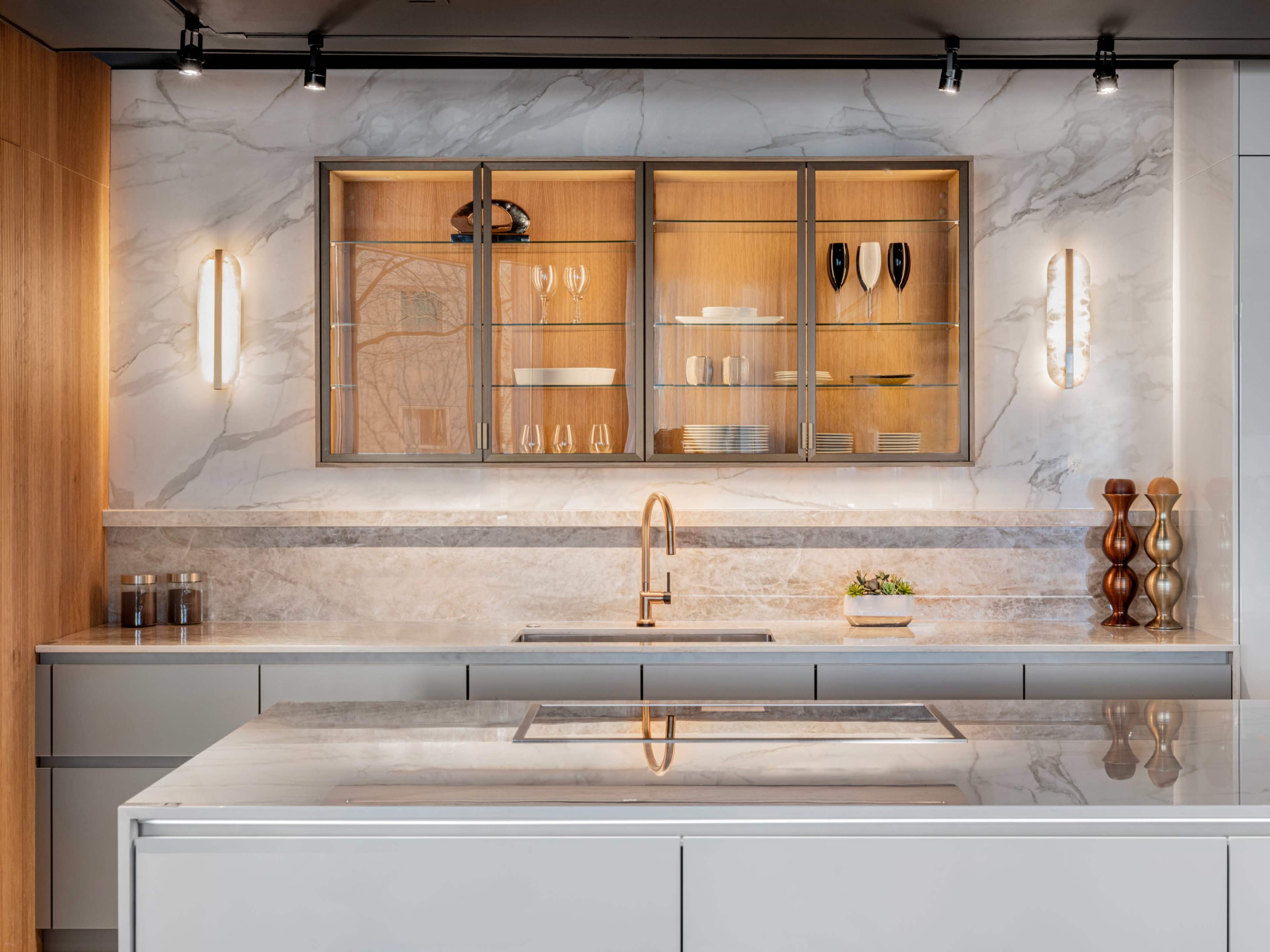 A modern kitchen features a marble countertop, sleek cabinets, and glass-fronted shelves displaying dishware.