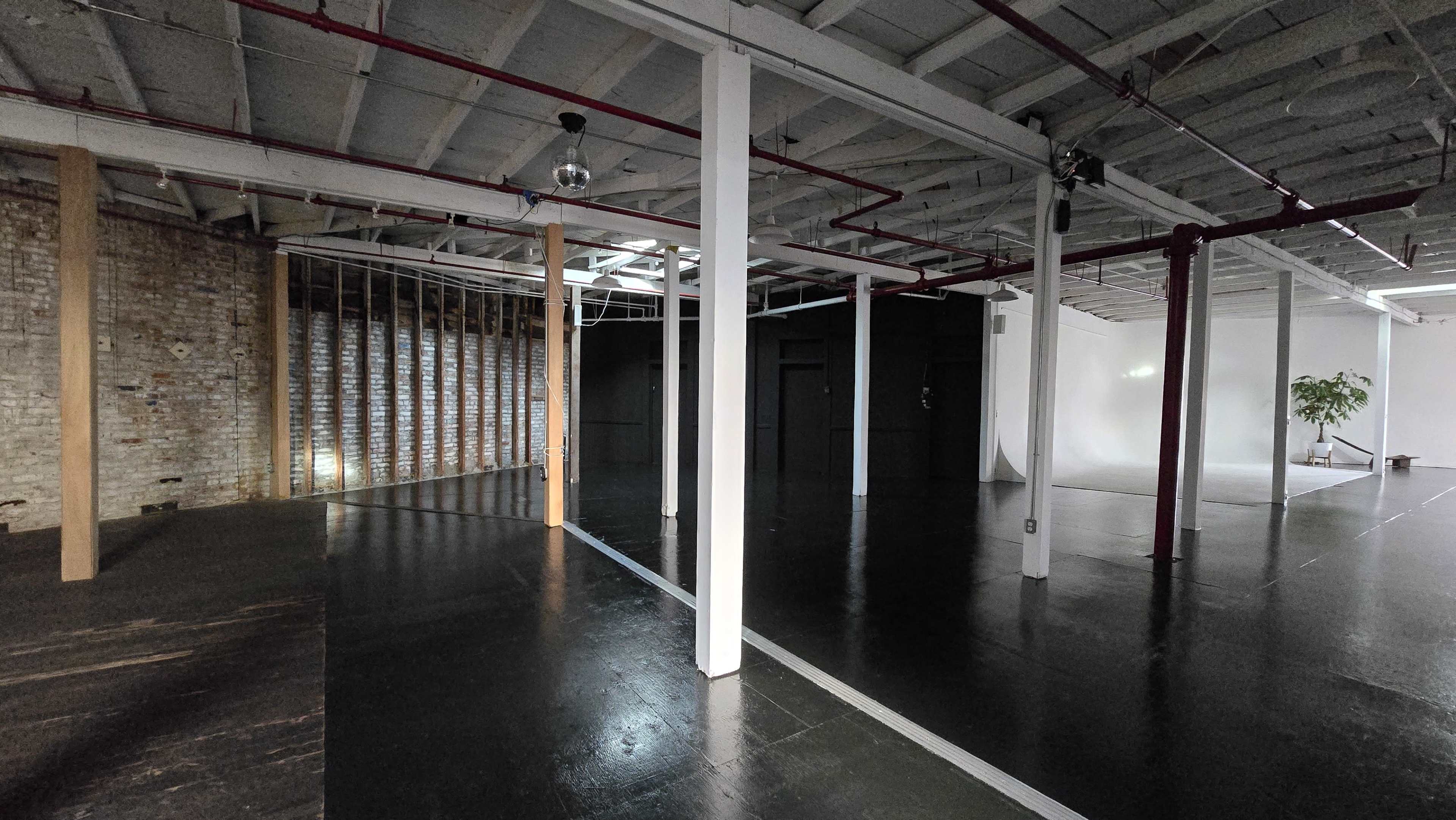 The image shows an empty industrial-style space with exposed brick walls, wooden beams, and distinct sections divided by pillars.