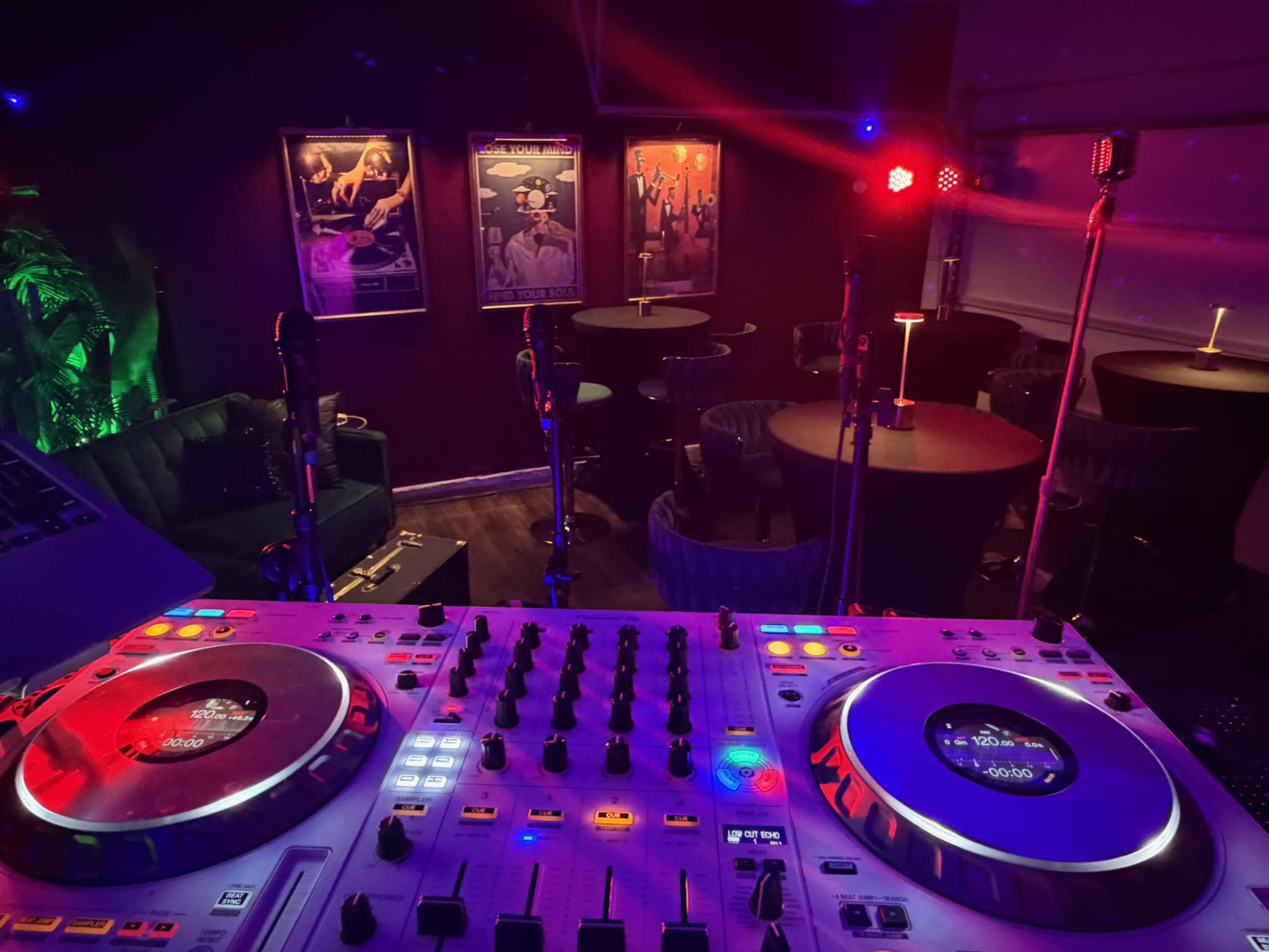 A DJ setup with turntables and mixers is in the foreground, while a dimly lit lounge area with tables and chairs is visible in the background, adorned with framed posters.