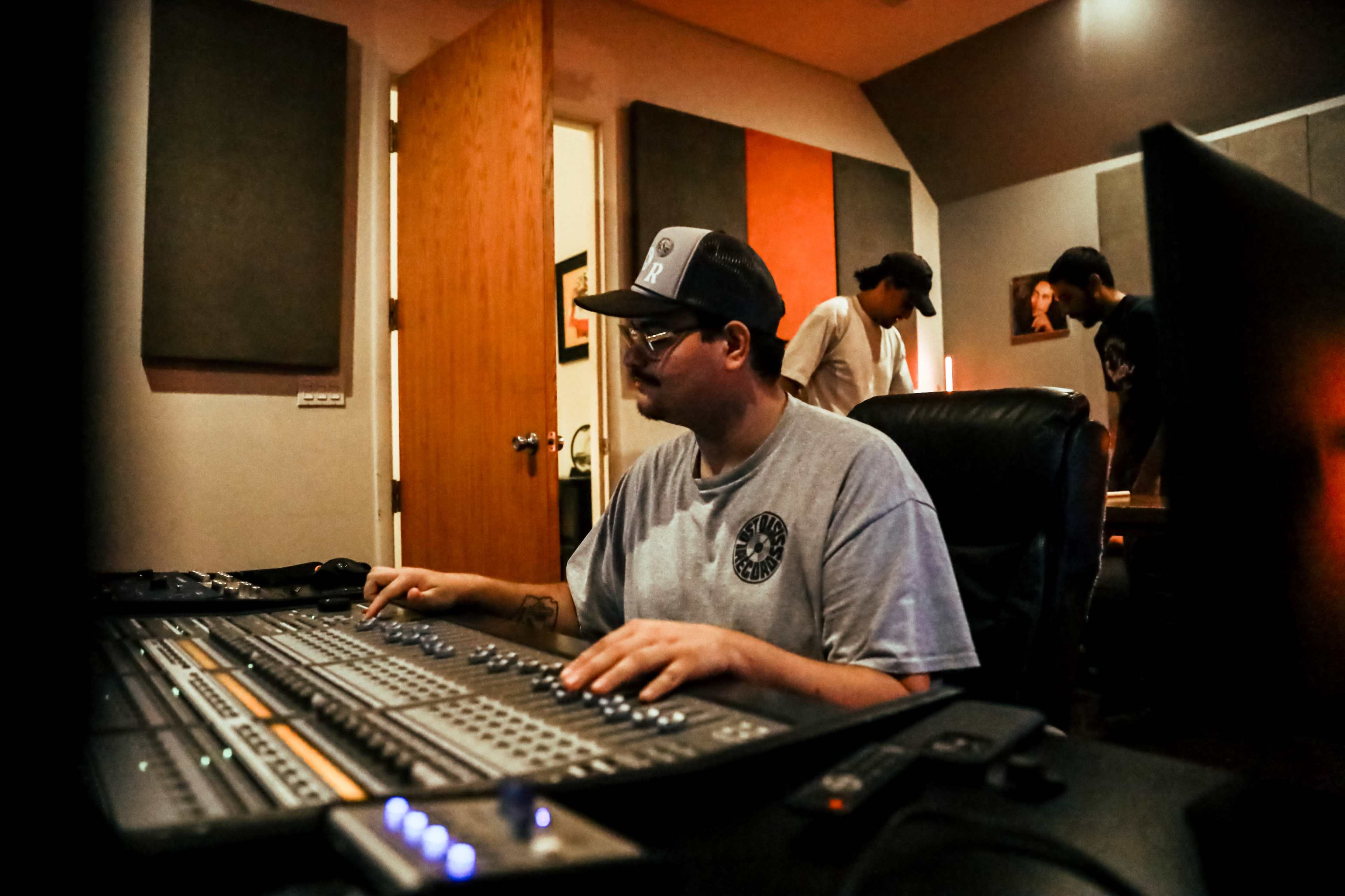 A man sits at a mixing console in a recording studio while two others work in the background.