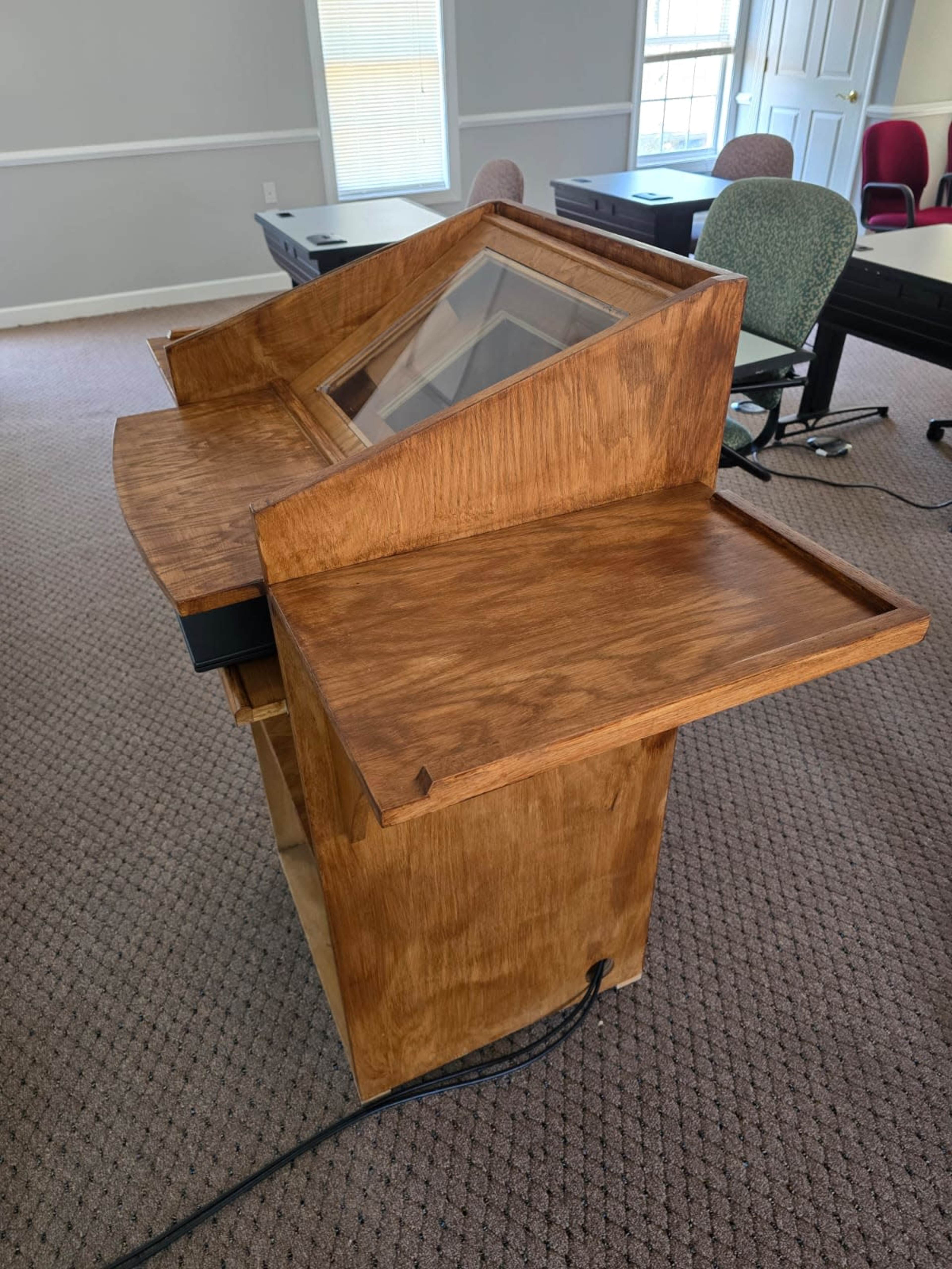 The image shows a wooden presentation podium with a slanted top and a drawer, positioned in a room with carpeted floors and tables.