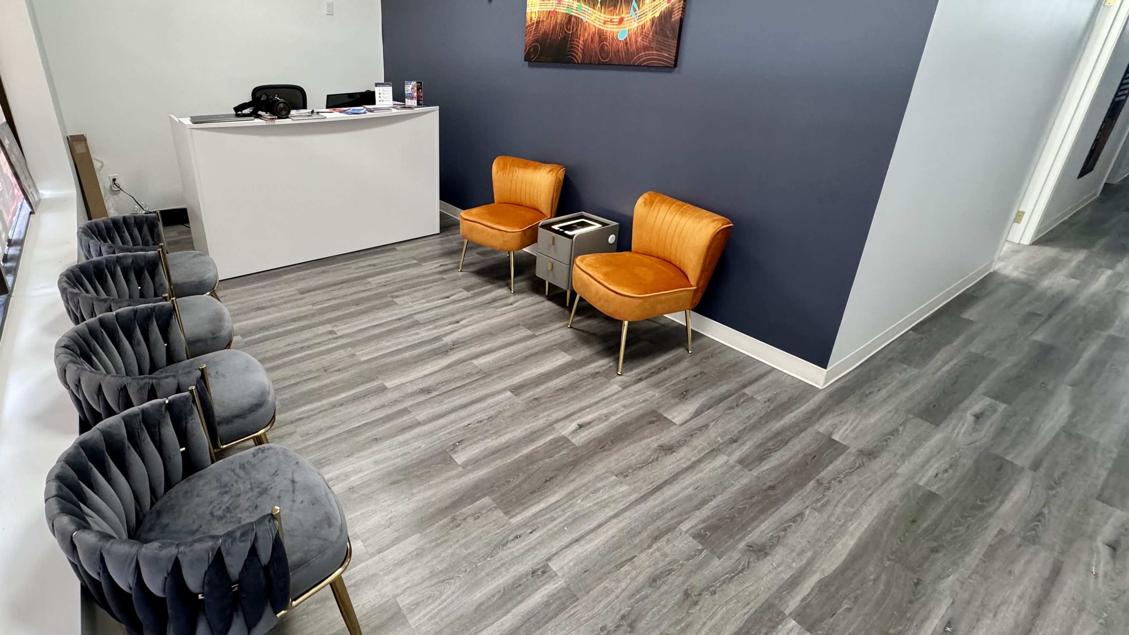 A waiting area with two orange chairs and several gray chairs arranged around a small table on a gray wooden floor.