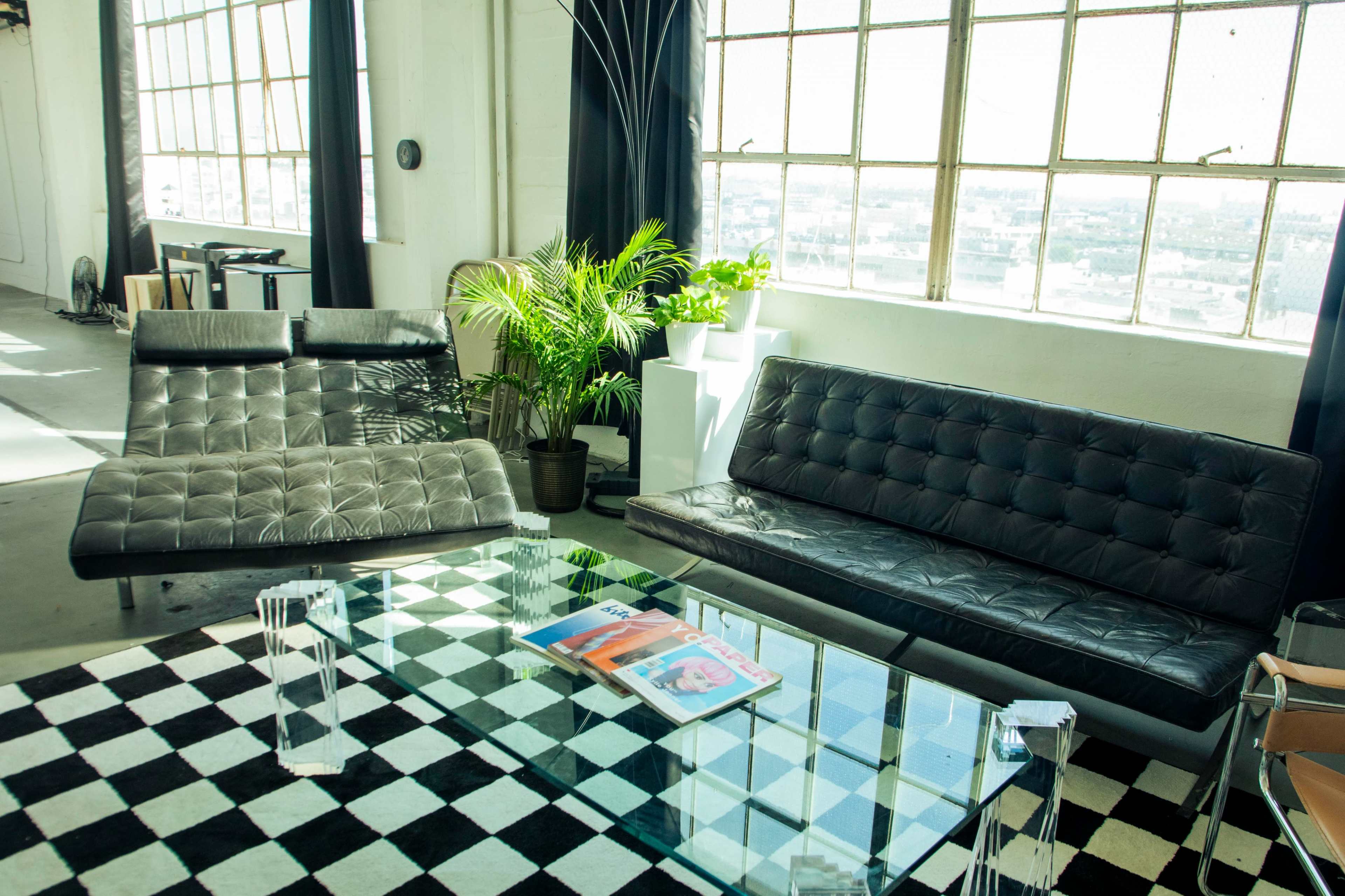 The image shows a modern interior with a glass coffee table, a black and white checkered rug, two sleek sofas, and large windows letting in natural light.
