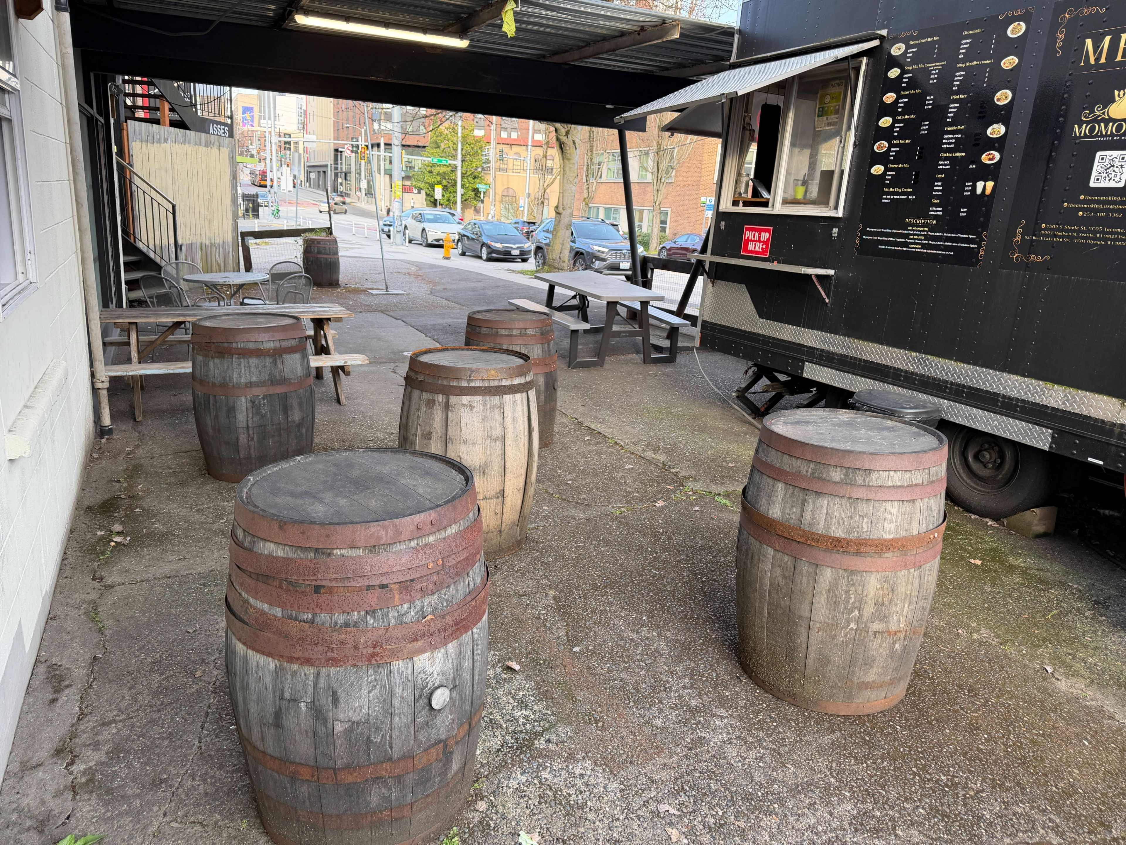 The image shows a food truck parked beside several wooden barrels arranged as seating in a paved outdoor area.
