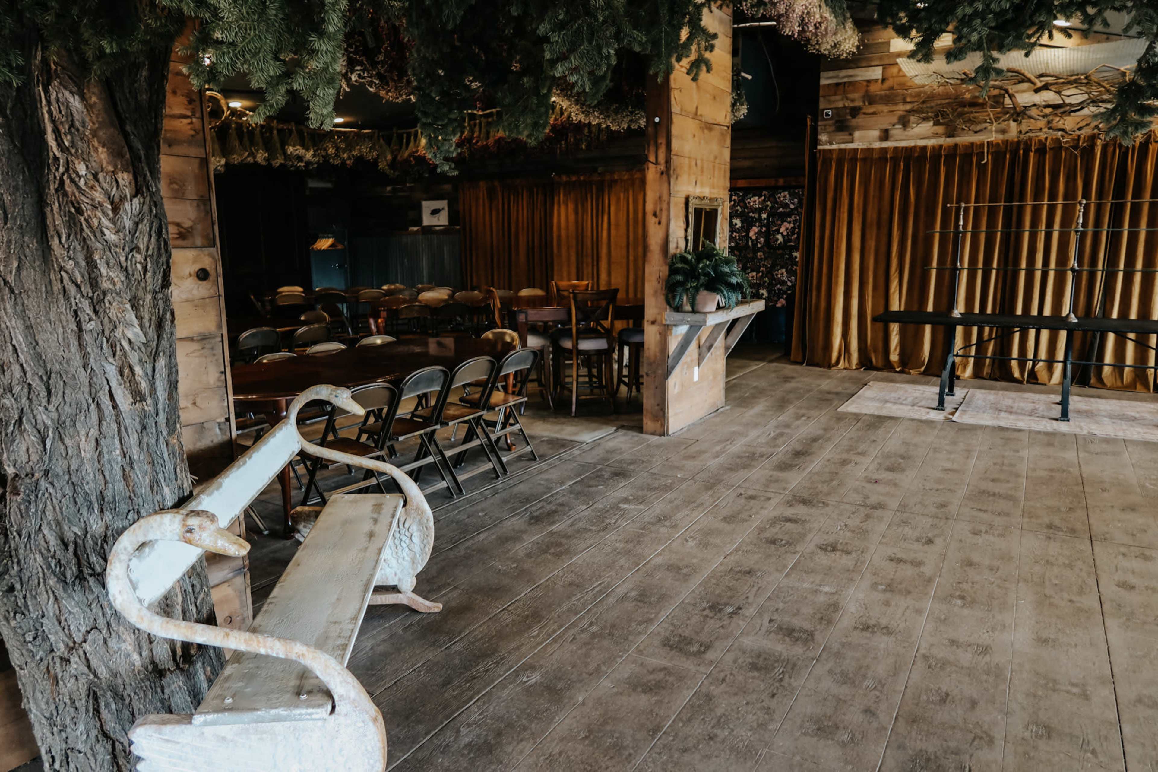 The image shows an interior room with wooden flooring, featuring a bench in the foreground, tables and chairs arranged for seating, and a backdrop of curtains and decor.