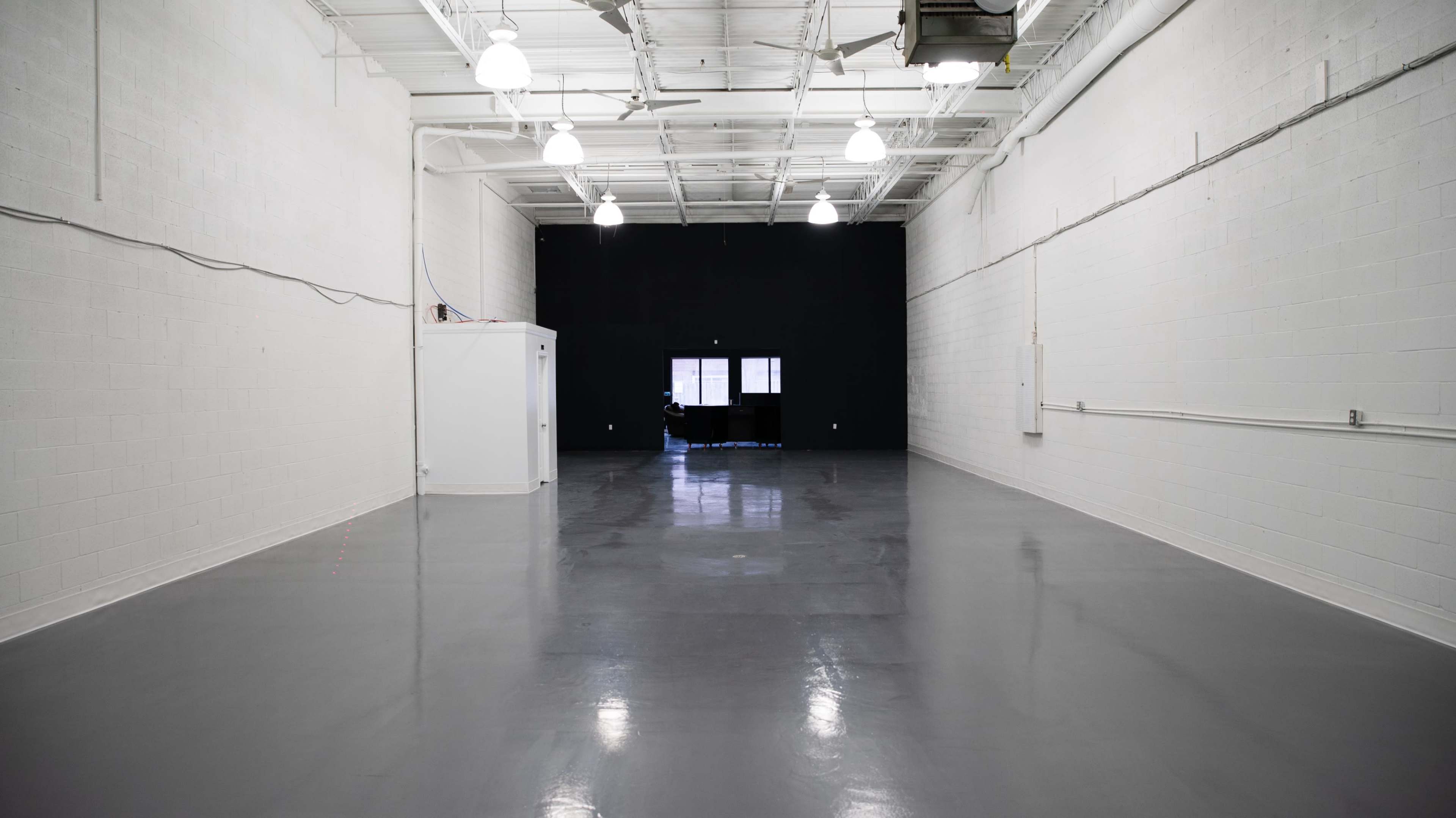 A spacious, empty room with gray floors, white walls, and a black wall at the far end, featuring a large window.