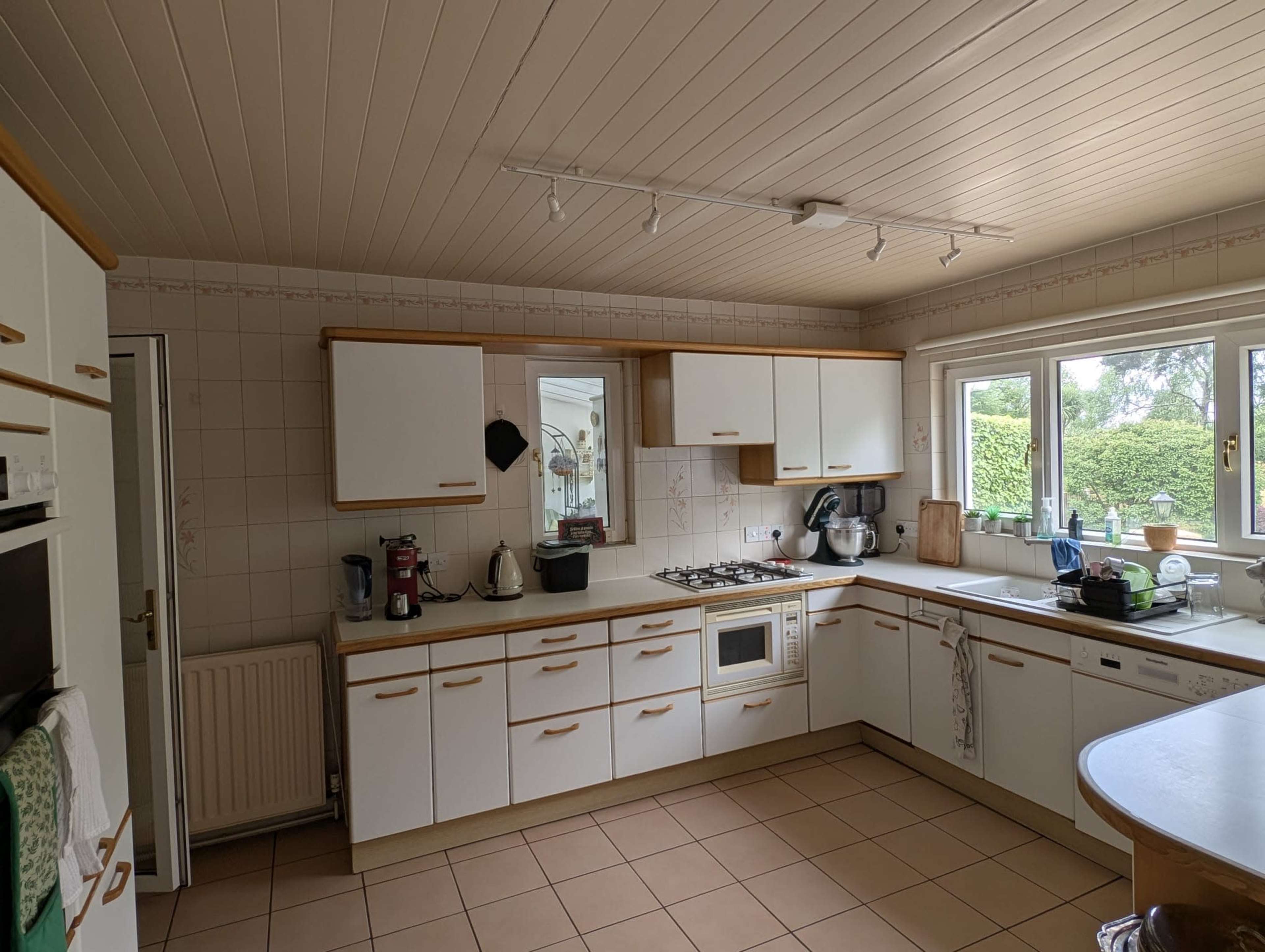 The image shows a spacious kitchen with white cabinetry, a tiled floor, and a window overlooking a garden.