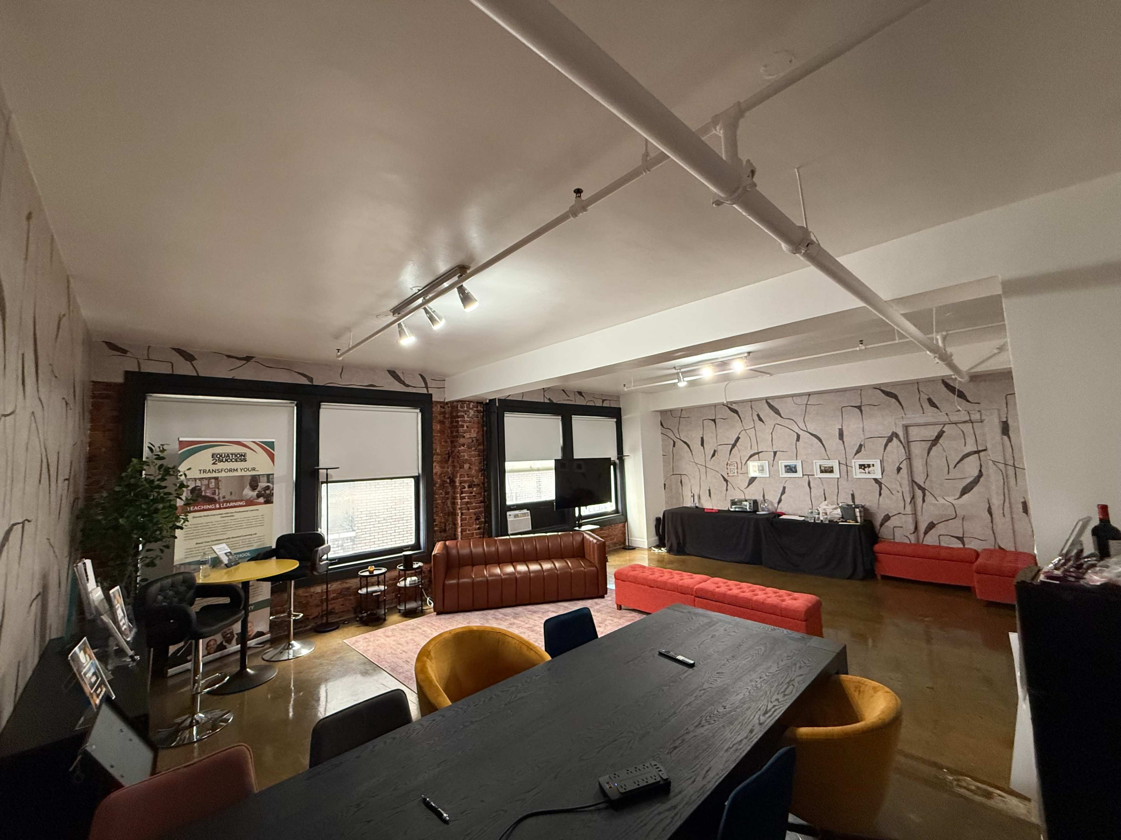 The image shows a modern office space featuring a long black table, colorful chairs, two seating areas with a brown leather couch and red sofas, and a decorative wall with a textured design.