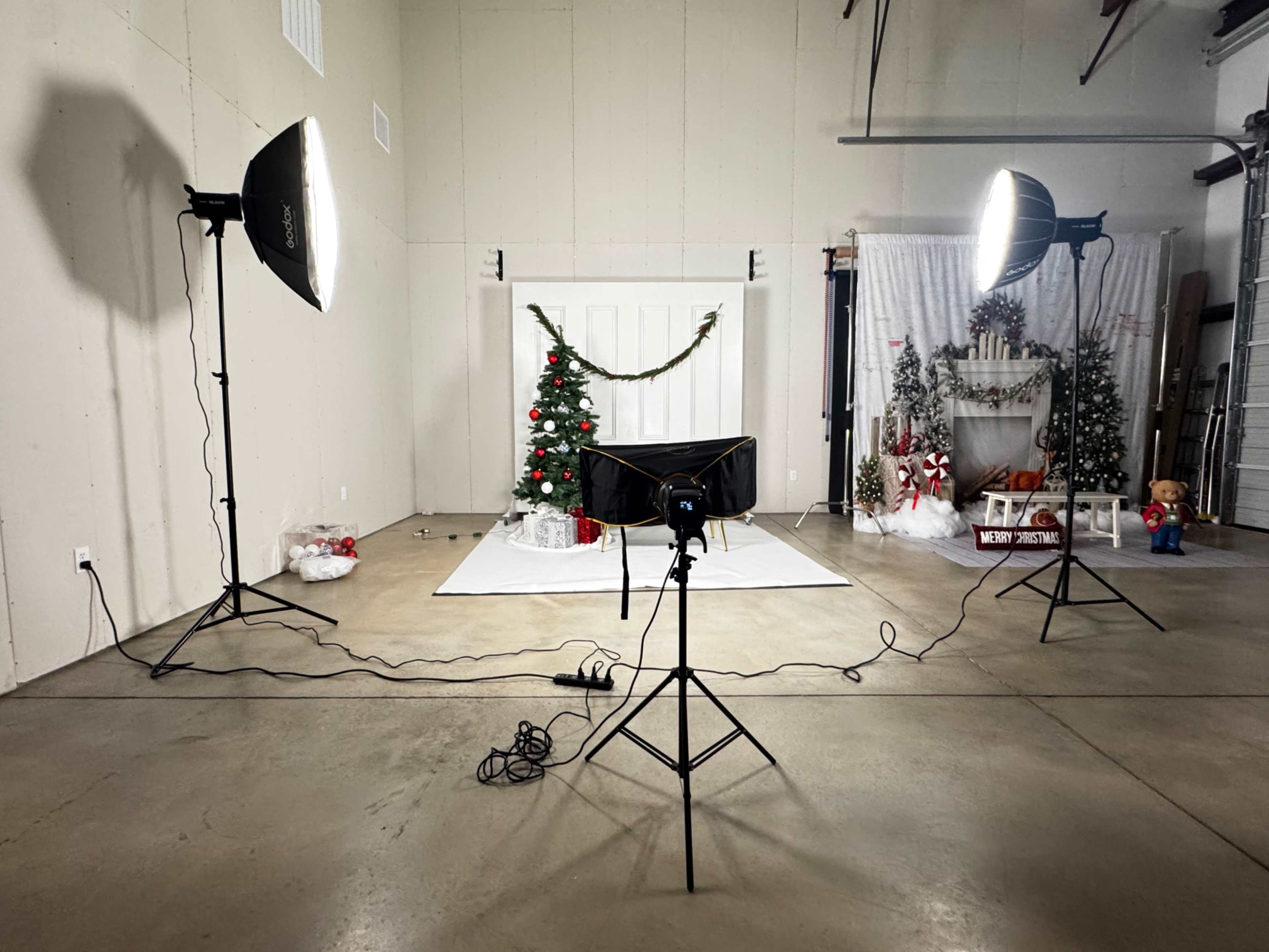 The image shows a studio setup for a Christmas-themed photo shoot, featuring a decorated tree, props, and multiple studio lights arranged around a white backdrop.