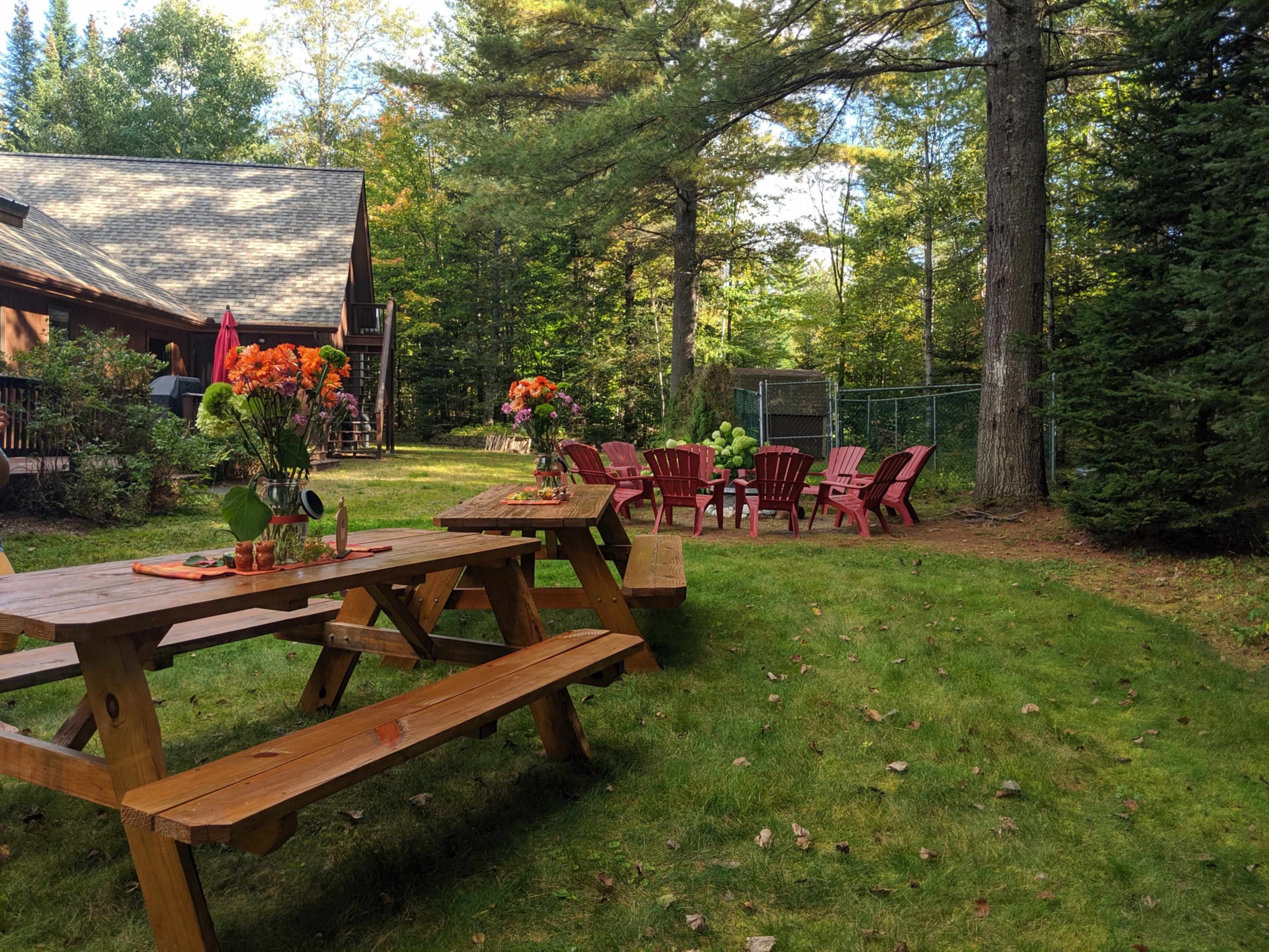 A backyard scene features wooden picnic tables and red Adirondack chairs arranged around a grassy area, with vibrant flower arrangements on the tables.