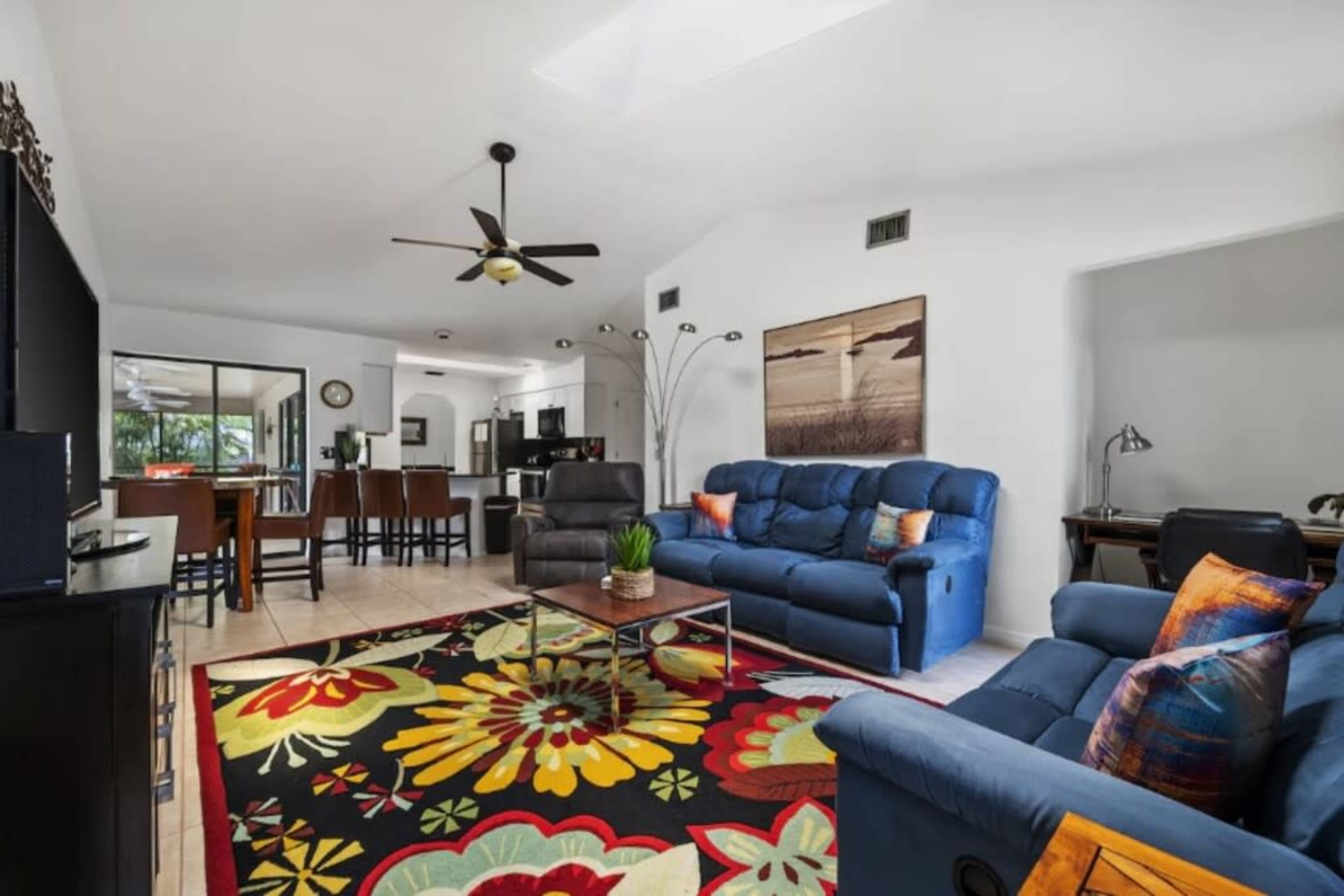The image shows a living room with two blue sofas, a coffee table on a colorful area rug, and a dining area visible in the background.