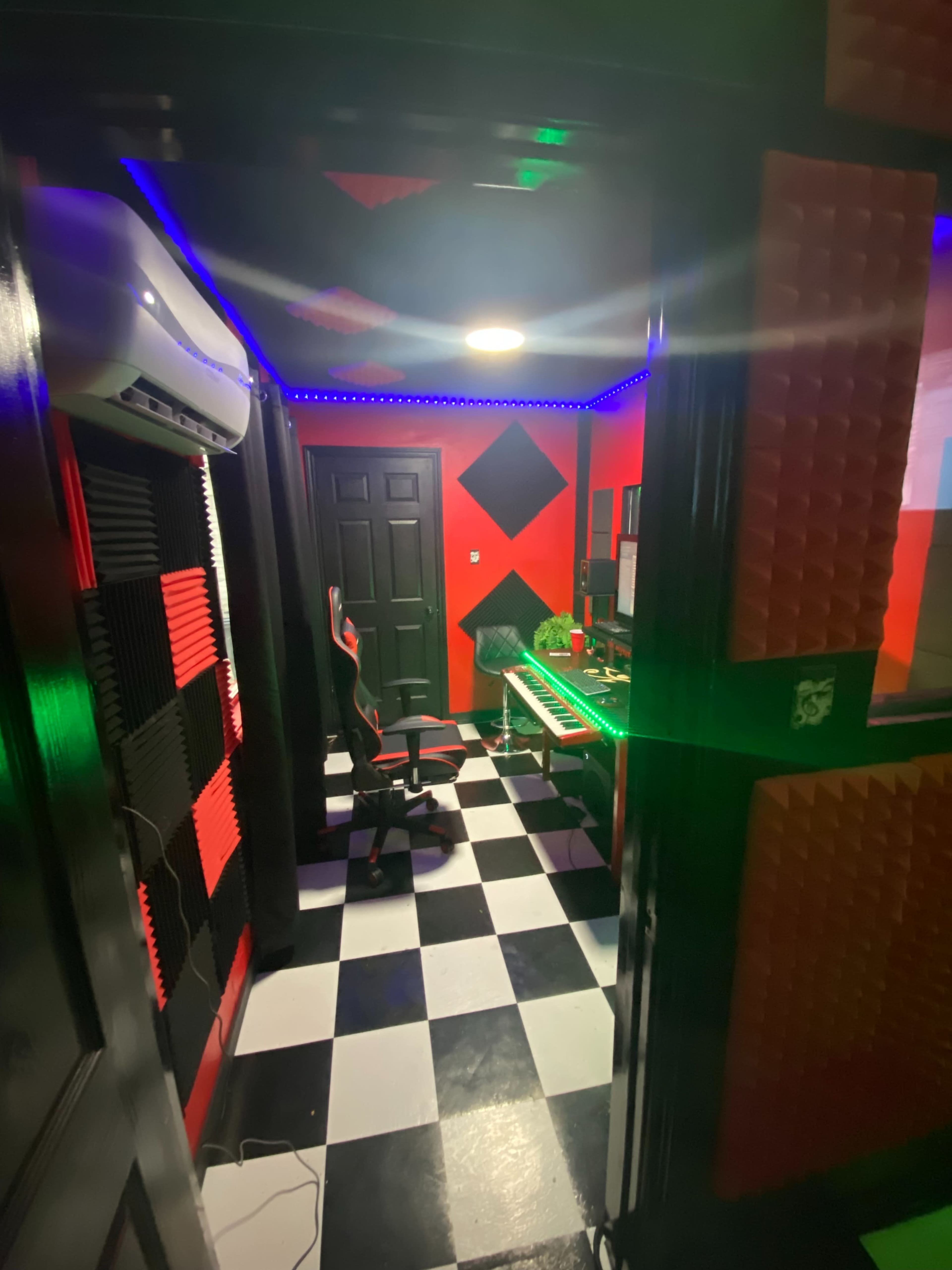 A soundproofed room with red and black walls, a checkered floor, an office chair, and a desk equipped with computer equipment and LED lighting.