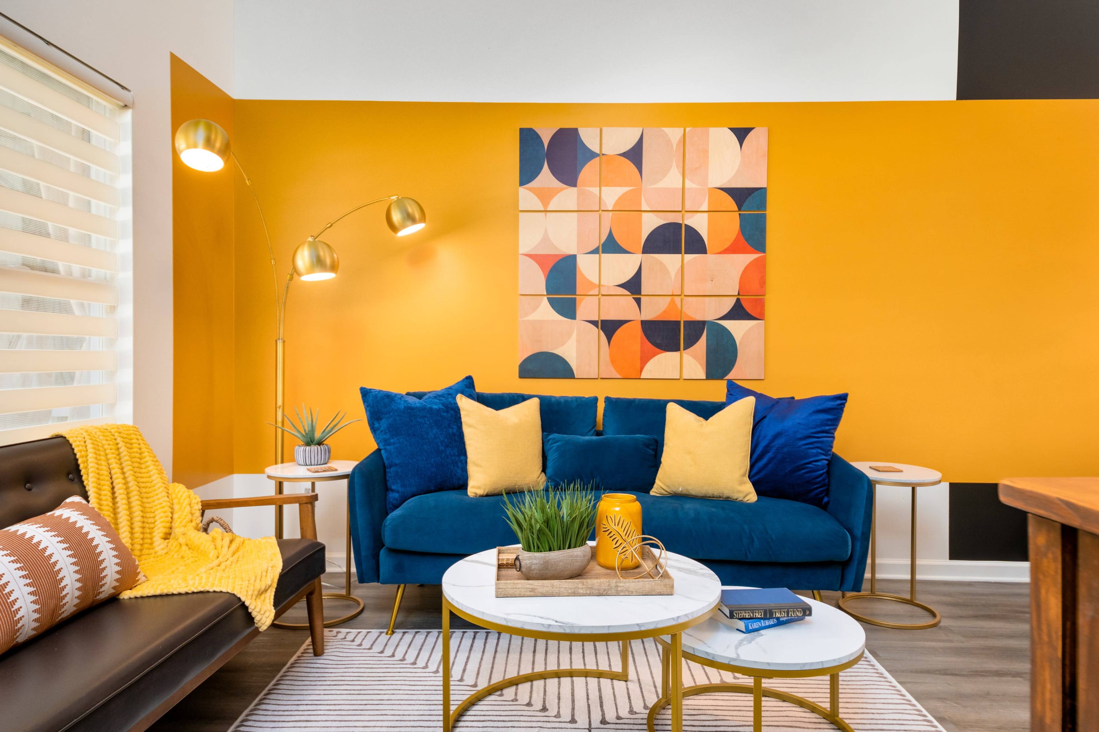 A modern living room features a blue velvet sofa with yellow and patterned cushions, a rounded coffee table, and a vibrant orange wall with abstract art.