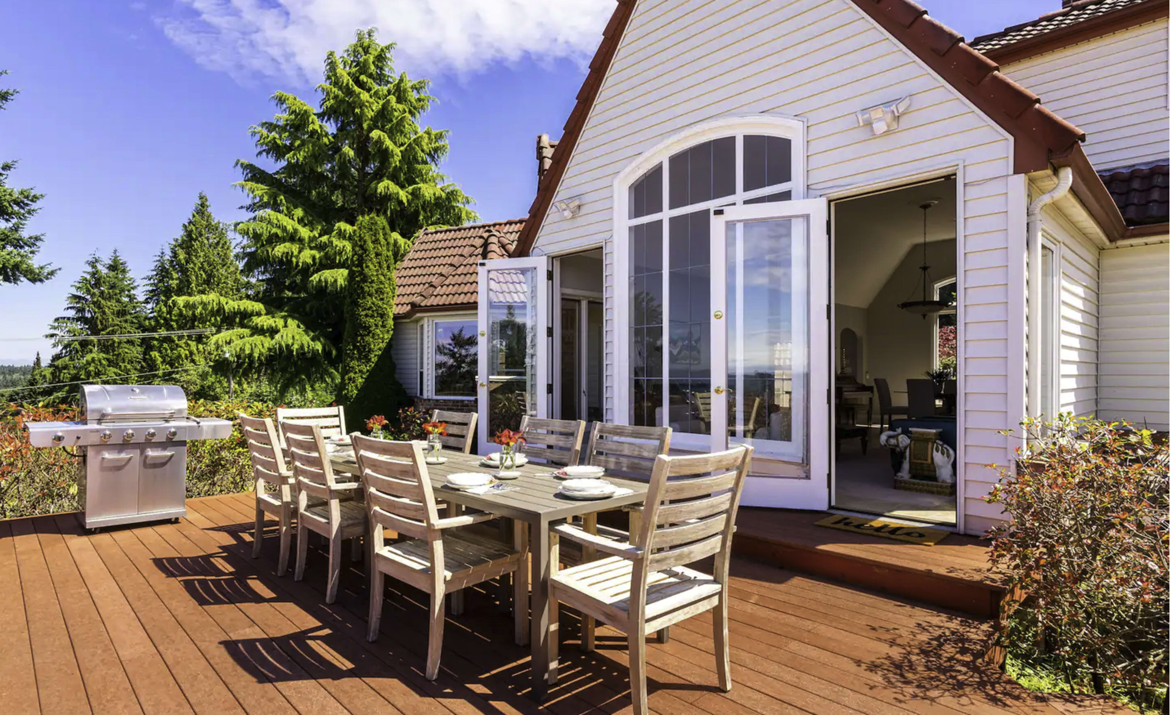 A wooden deck with a dining table and chairs is set up outside a house, featuring a grill and a view of trees and a blue sky.