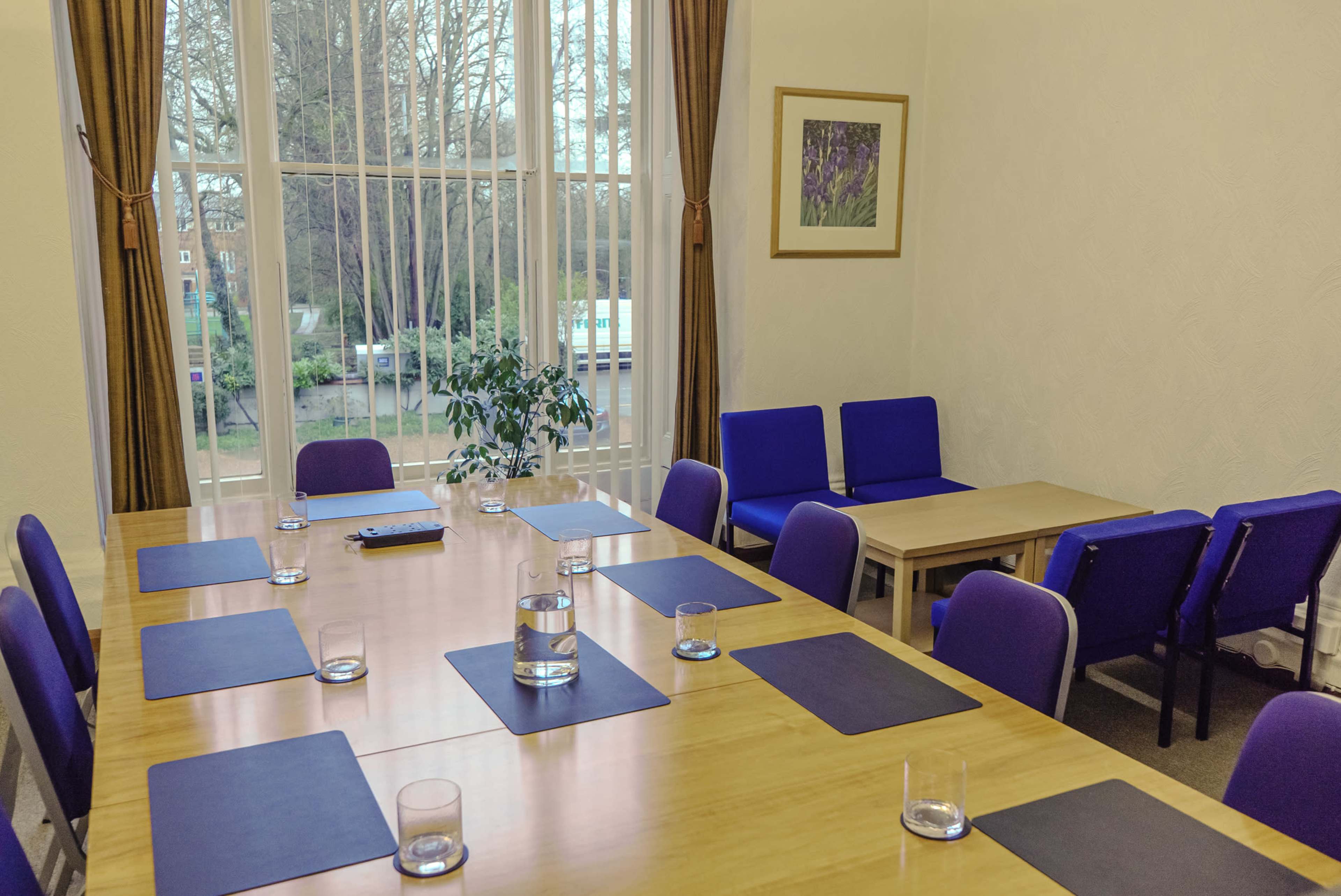 The image shows a well-lit meeting room with a long wooden table set with glasses and placemats, surrounded by blue chairs, and featuring large windows with vertical blinds.