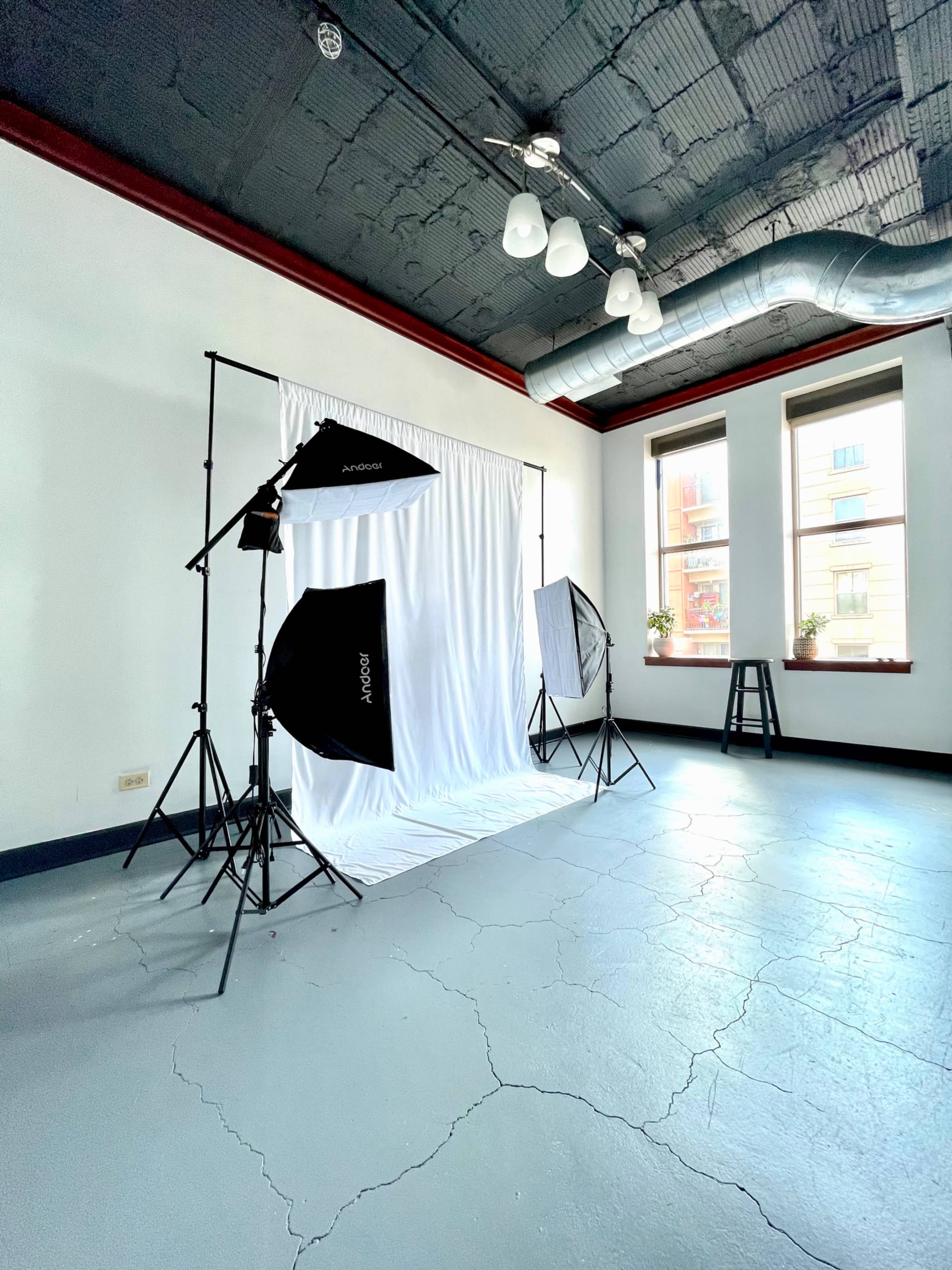 A photography studio setup with a white backdrop, lighting equipment, and a stool in a room with large windows.