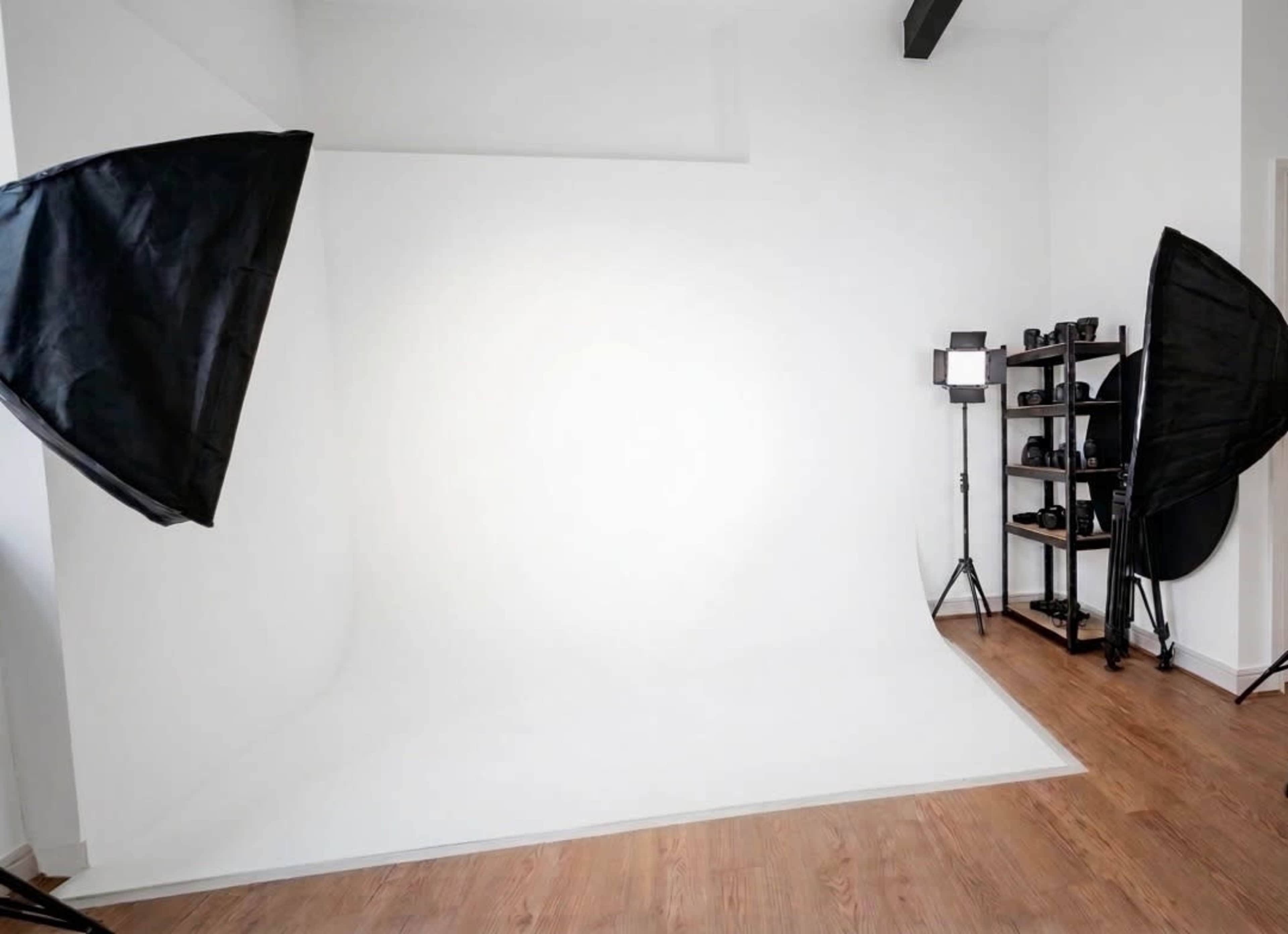 The image shows a photography studio with a white backdrop, two softbox lights on either side, and a shelf holding camera equipment in the corner.