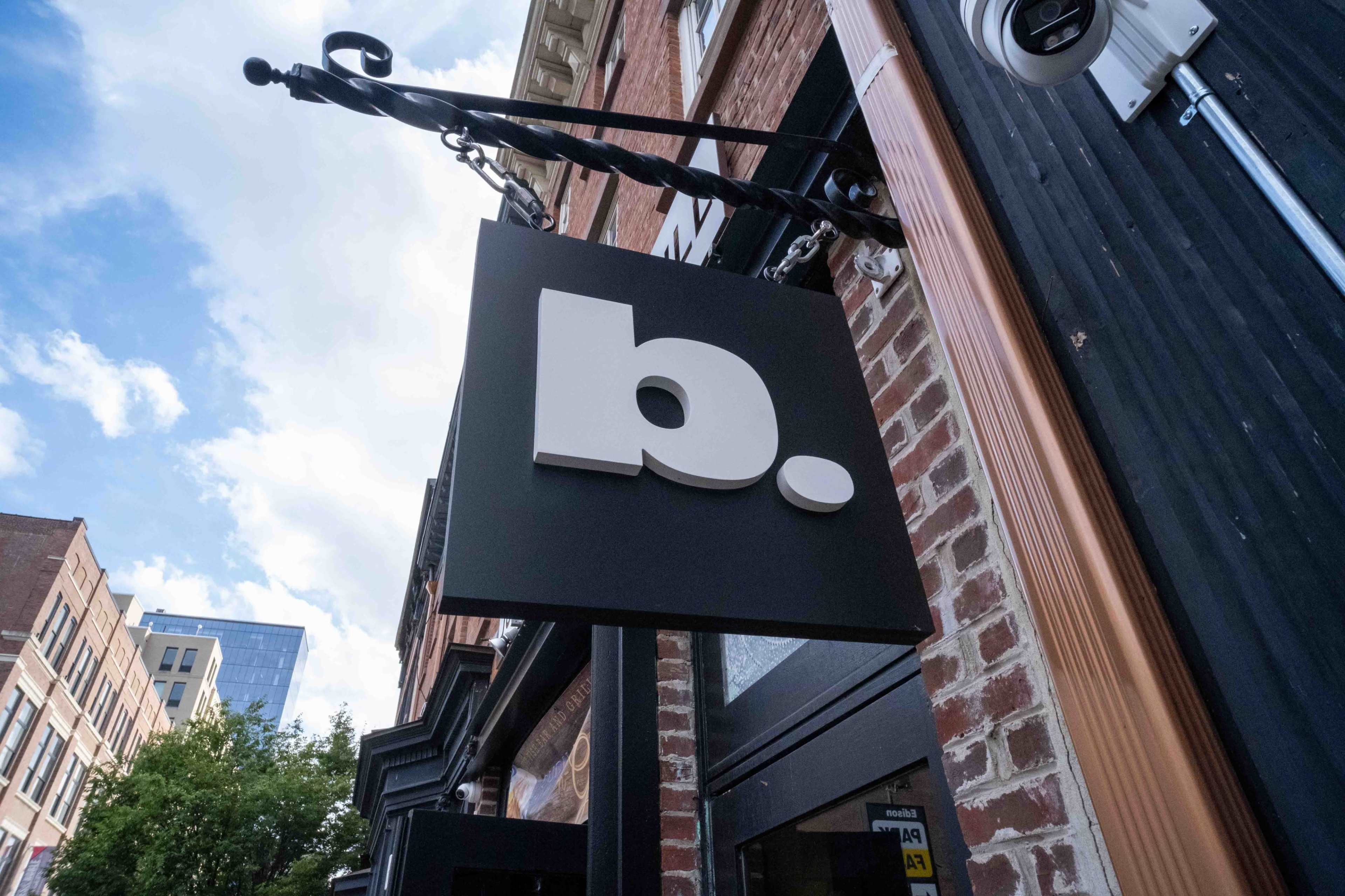 A large black sign with a white letter "b." hangs above a building entrance in an urban setting.