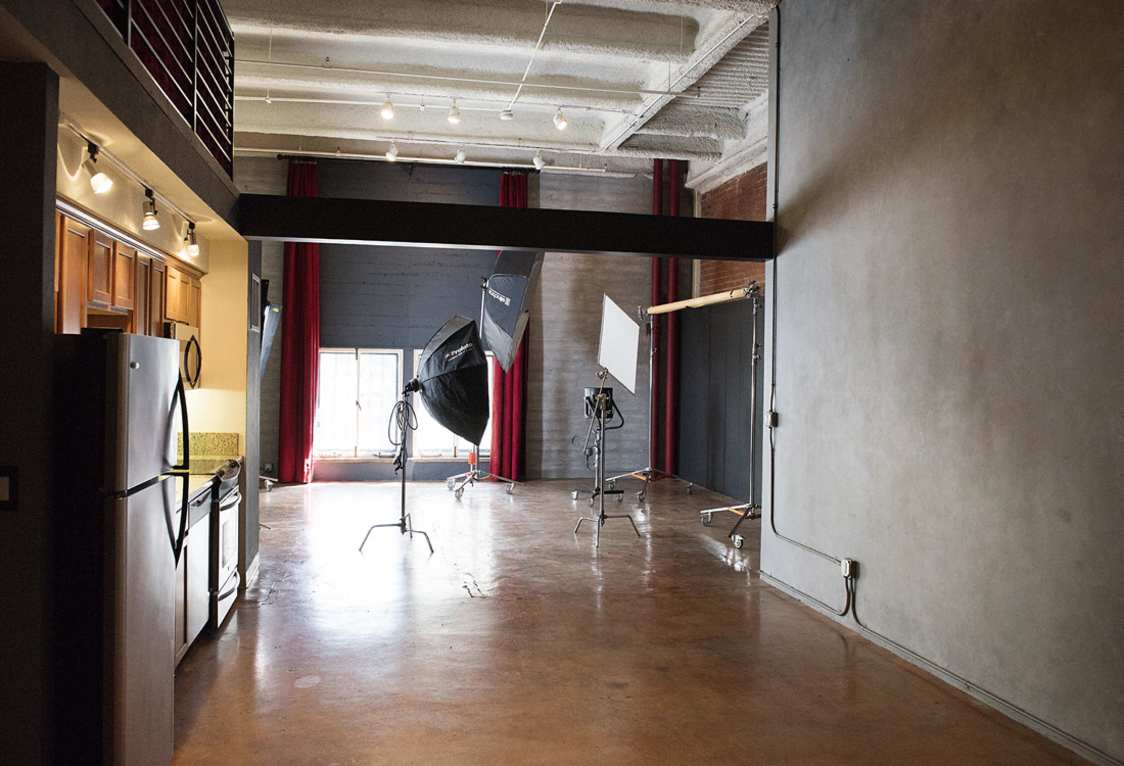 The image shows an industrial-style interior with a kitchen on the left and photography equipment set up on the right, including softboxes and a backdrop.