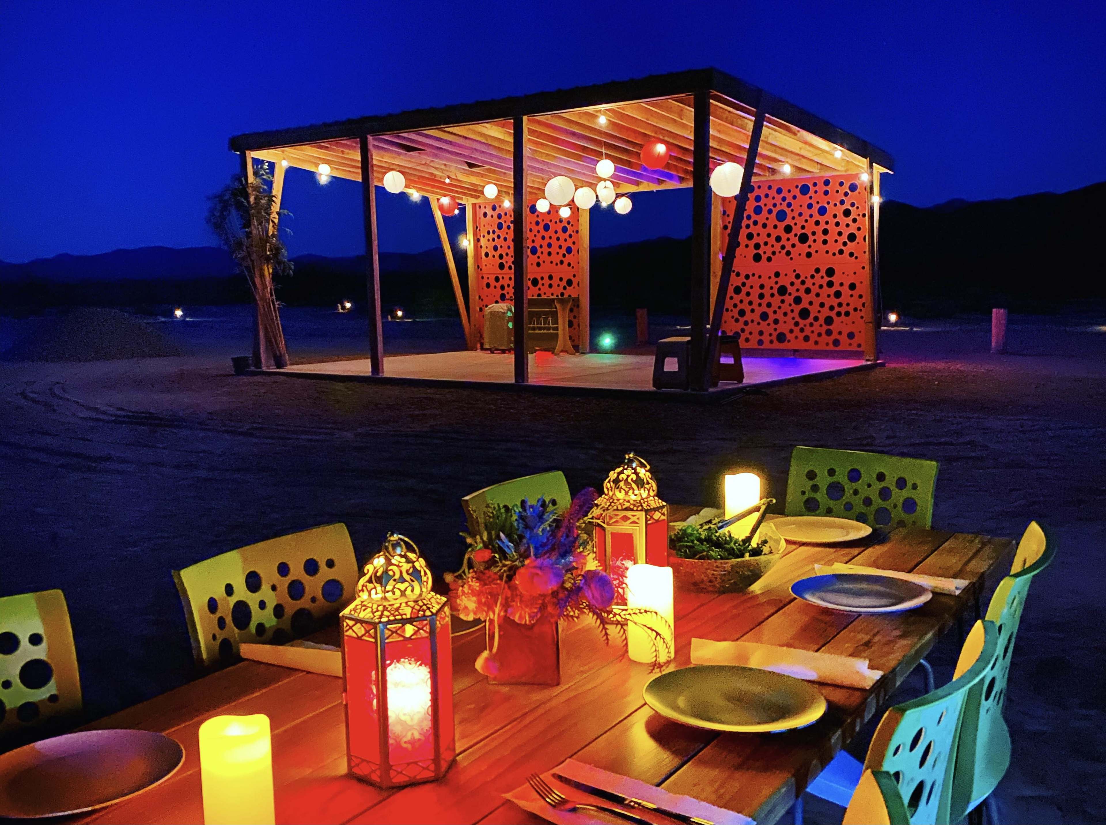 A wooden dining table with colorful chairs is set up outside a lit pavilion decorated with hanging lanterns and a vibrant backdrop under a night sky.
