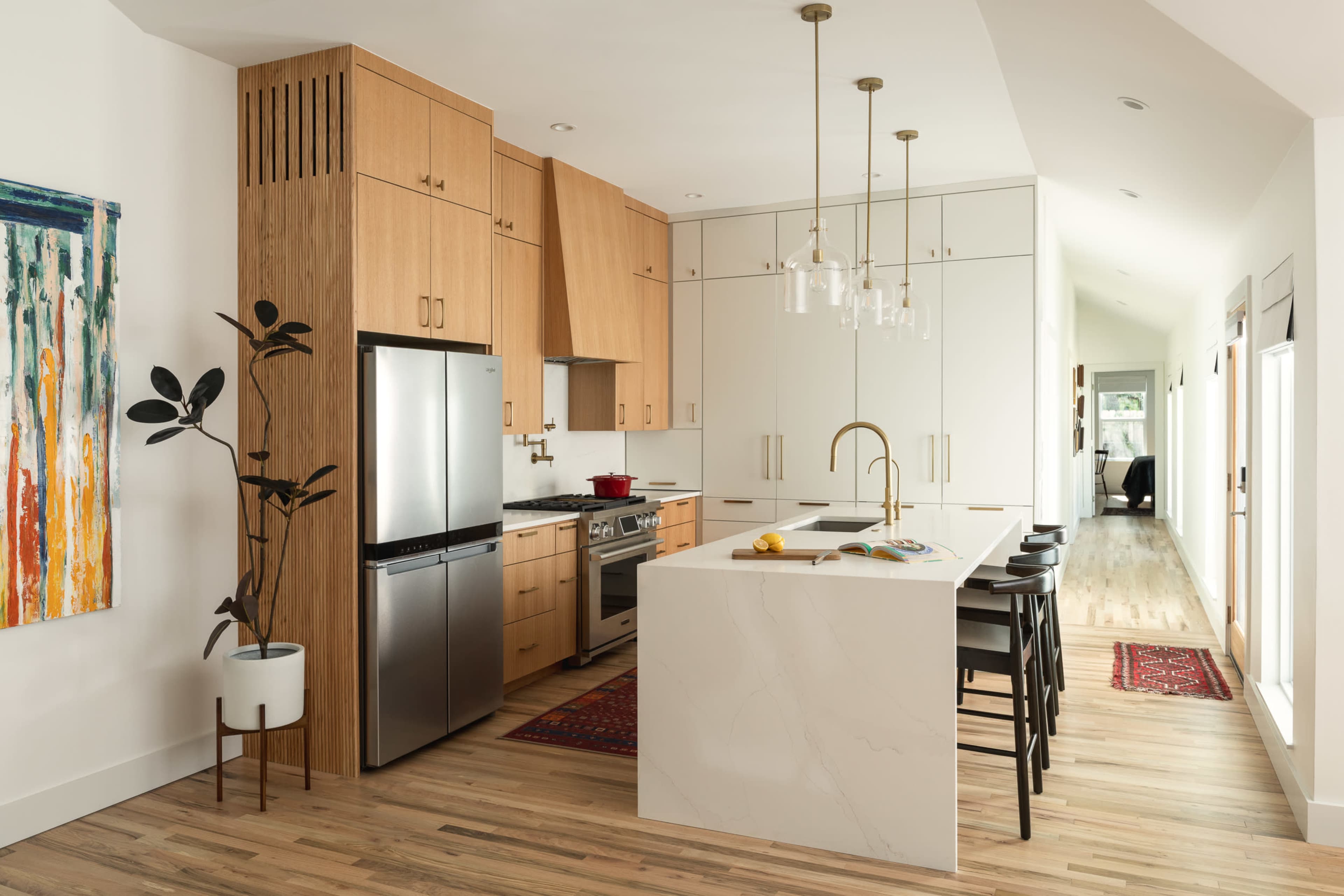 The image shows a modern kitchen featuring wooden cabinetry, stainless steel appliances, a large island with bar seating, and an adjacent hallway leading to other areas of the home.