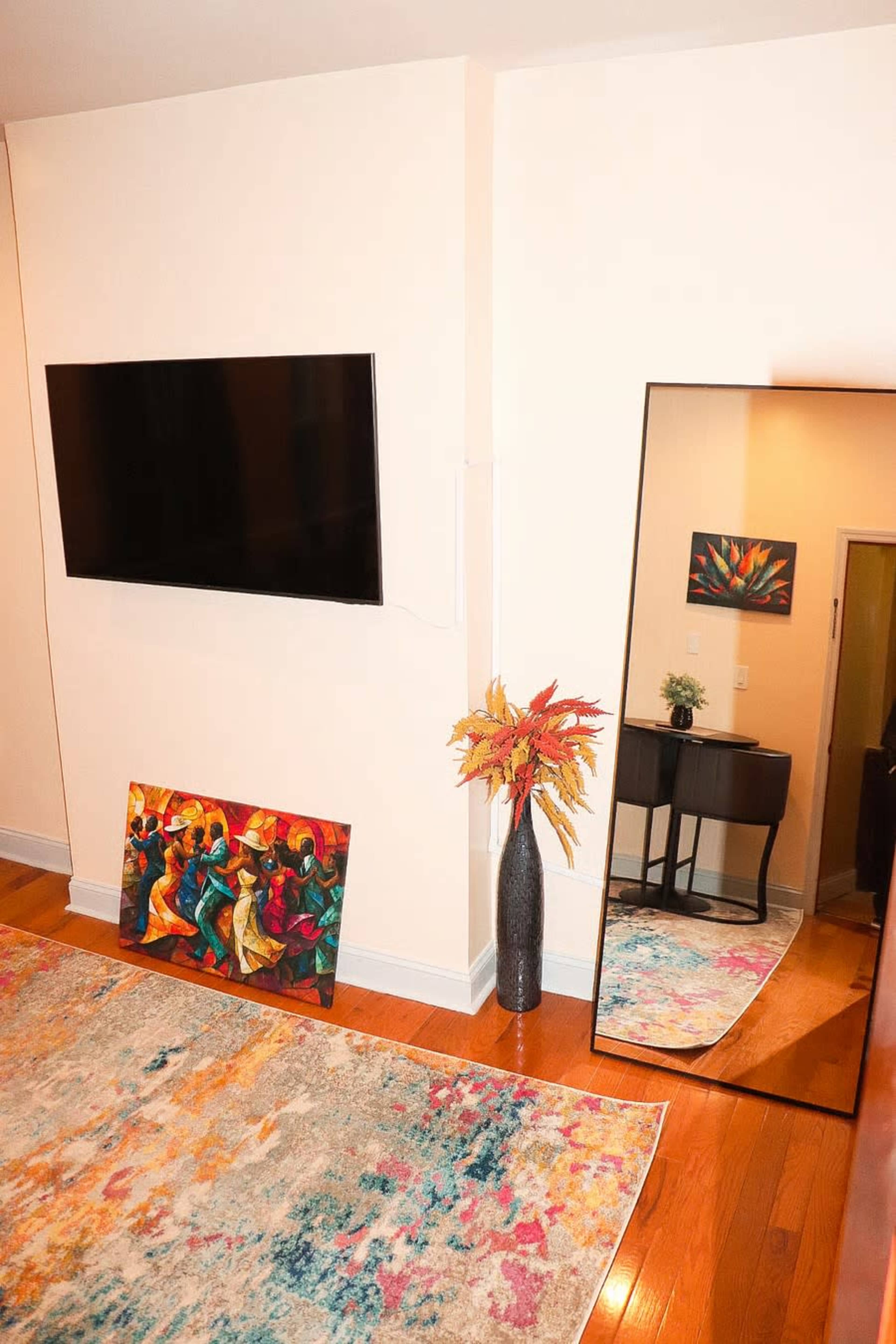 The image shows a modern living room with a mounted flat-screen TV, an artistic painting on the floor, a tall vase with decorative flowers, and a large mirror reflecting a hallway.