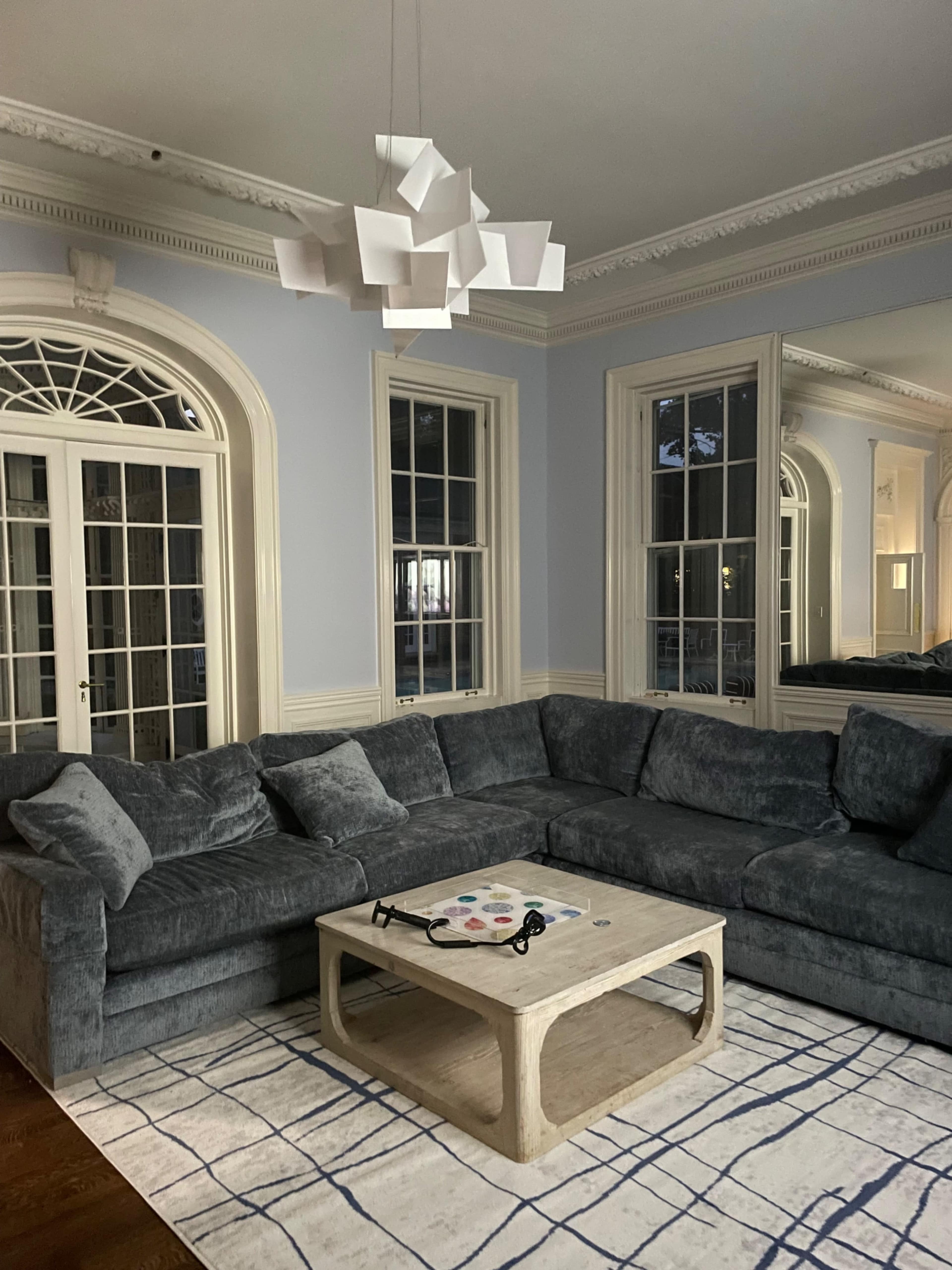 The image shows a living room with a sectional sofa, a coffee table, and large windows allowing natural light to enter the space.