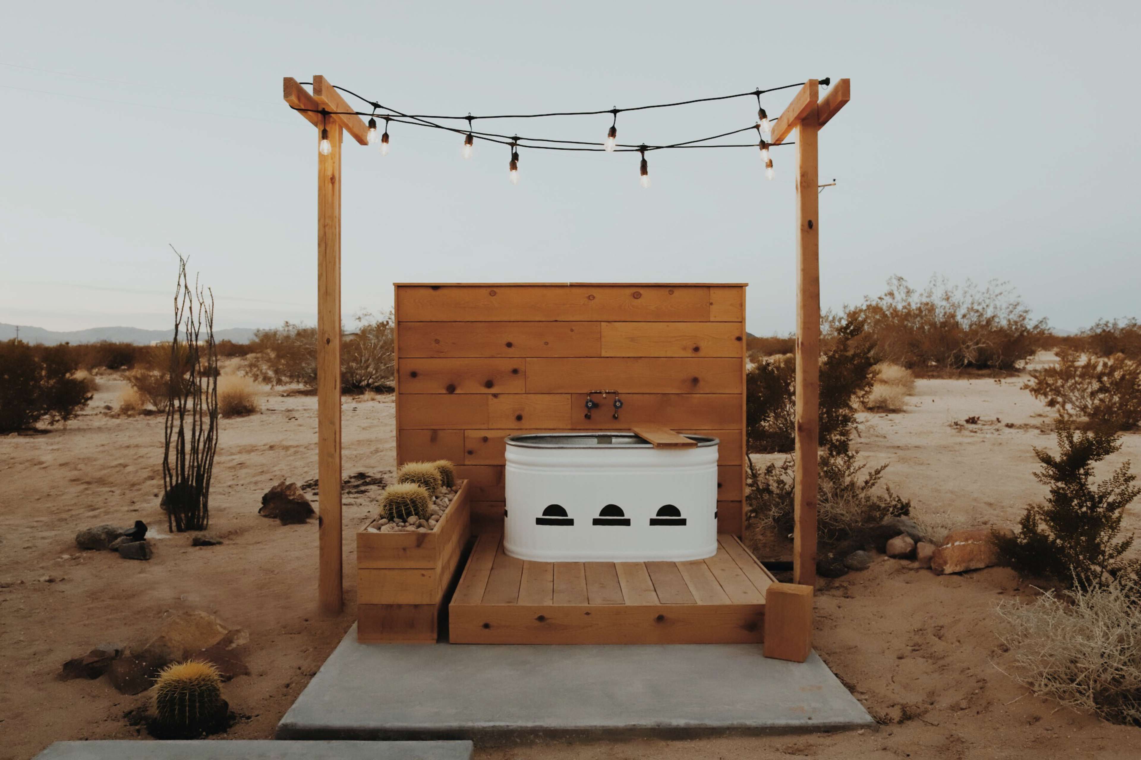 The image shows an outdoor hot tub enclosed by wooden decking and a rustic wooden wall, illuminated by string lights in a desert setting.