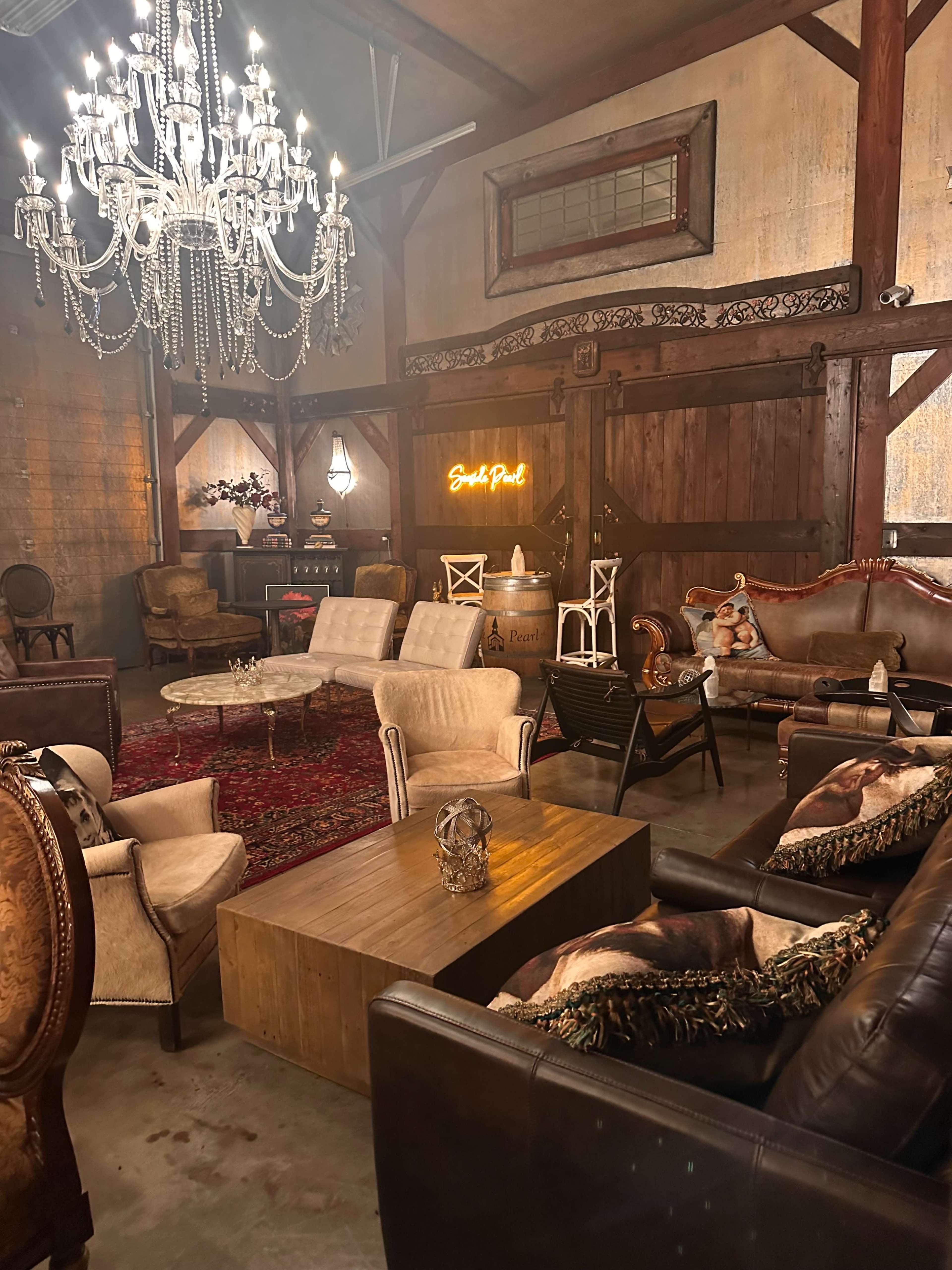 The image shows a rustic interior with various seating arrangements, including vintage sofas and chairs, under a large chandelier, featuring a neon sign that reads "Spark Bar."