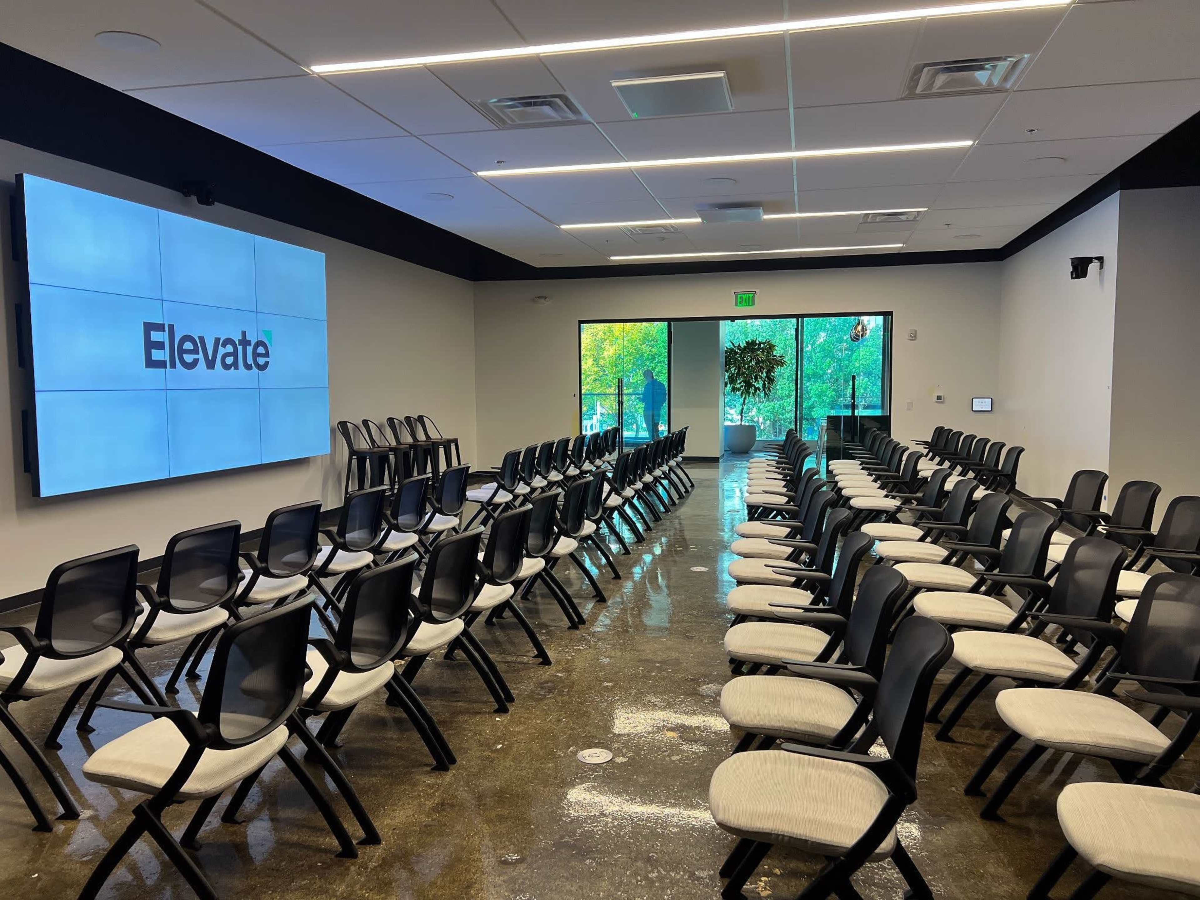 A modern conference room is set up with rows of black and white chairs facing a large screen displaying the word "Elevate."