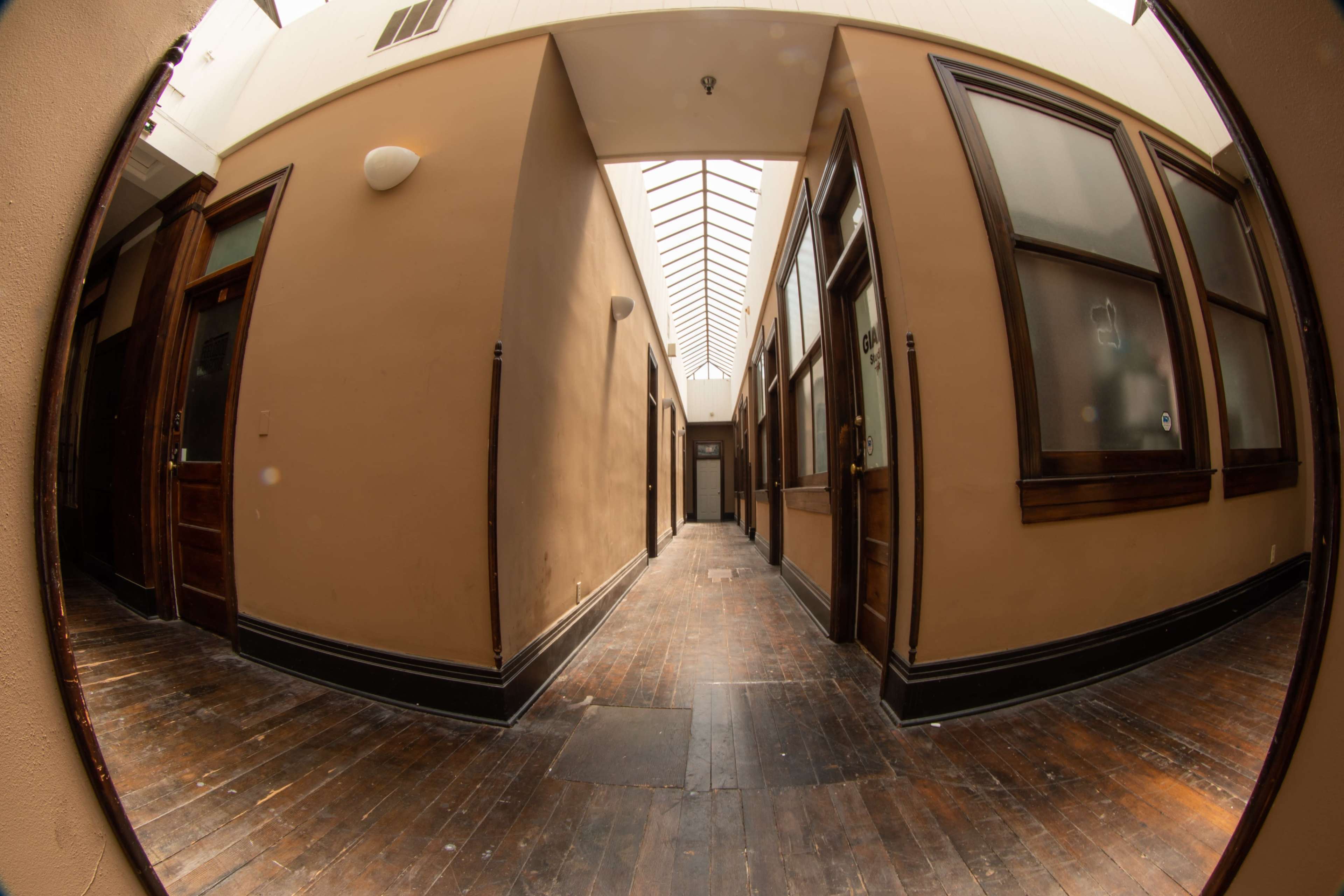 The image shows a narrow corridor with wooden floors and doors on either side, illuminated by a skylight above.