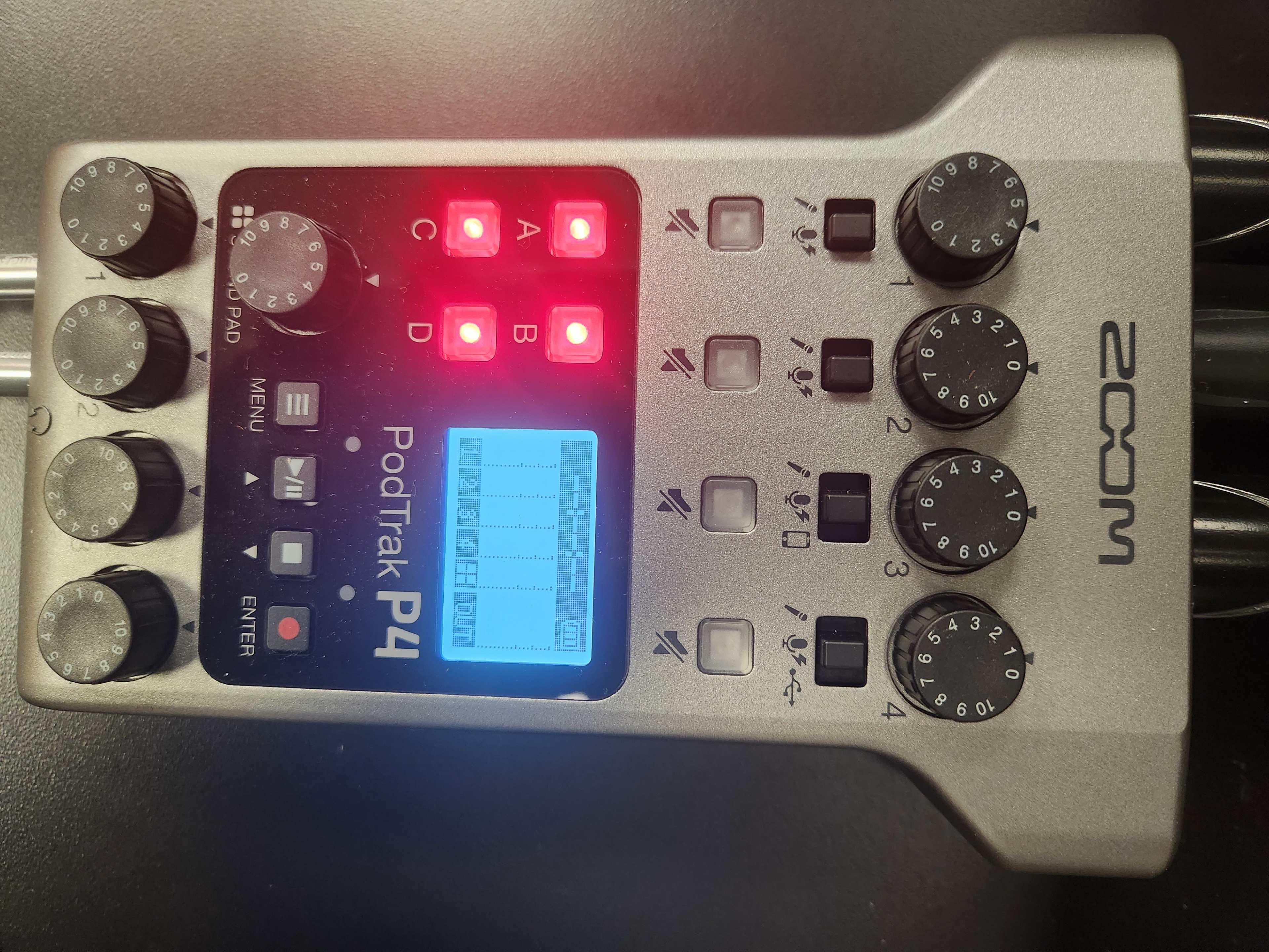 The image shows a Zoom PodTrak P4 audio recorder with various knobs, buttons, and an illuminated display.