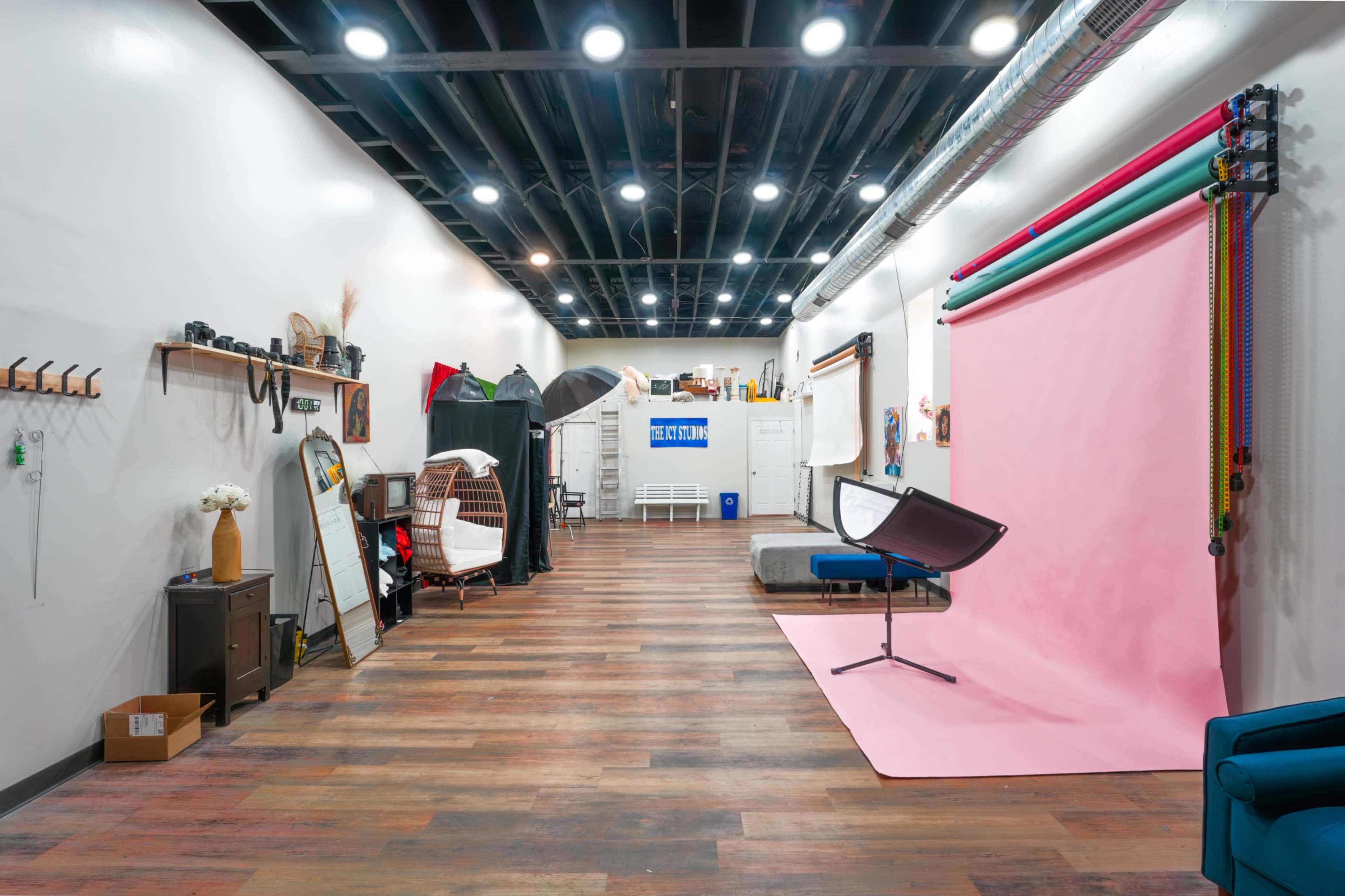 The image shows a spacious photography studio featuring a pink backdrop, various equipment, and wooden flooring.