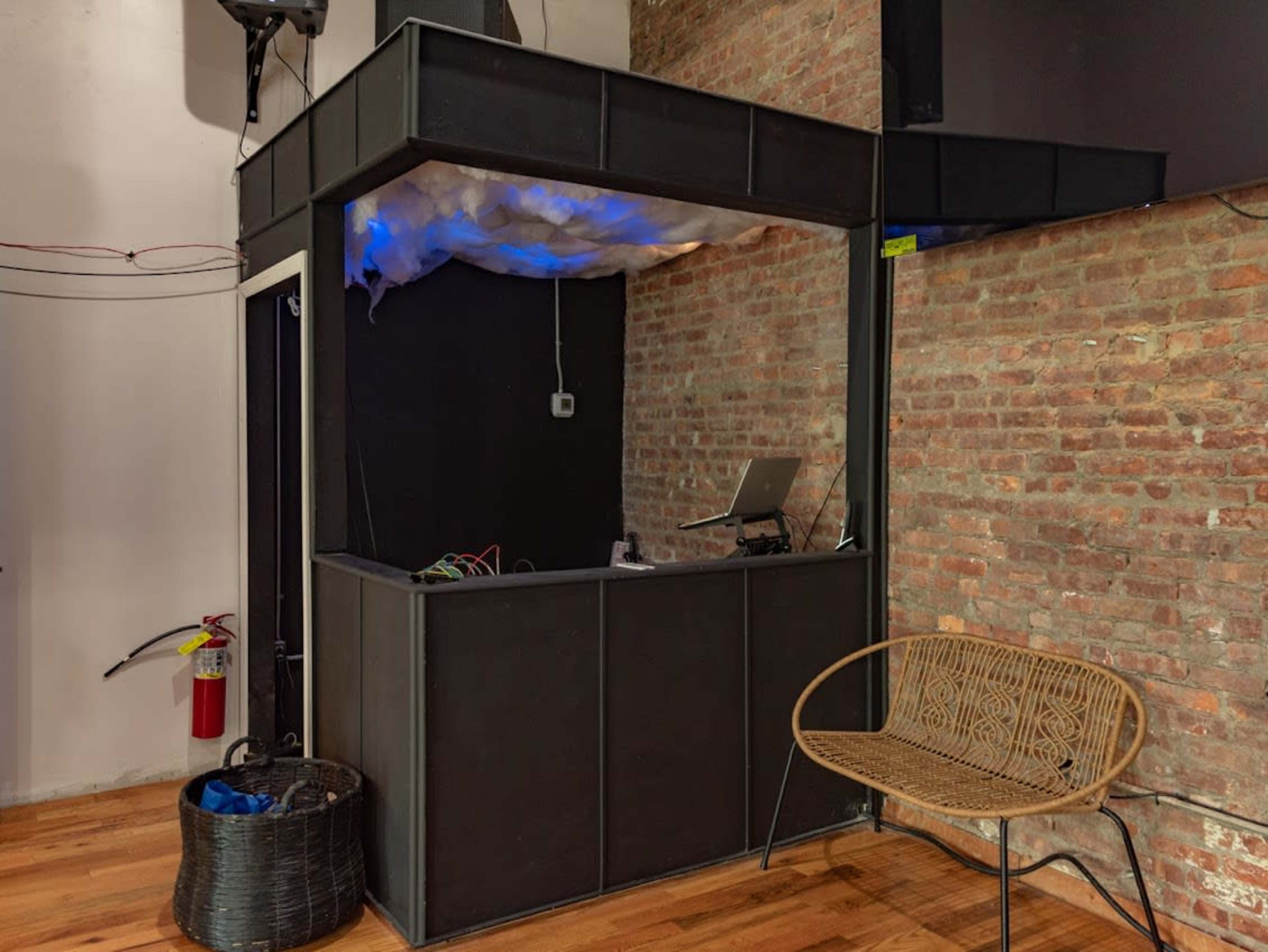 The image shows a black boxy structure with a small window and a desk holding a laptop, situated in a room with exposed brick walls and wooden flooring.