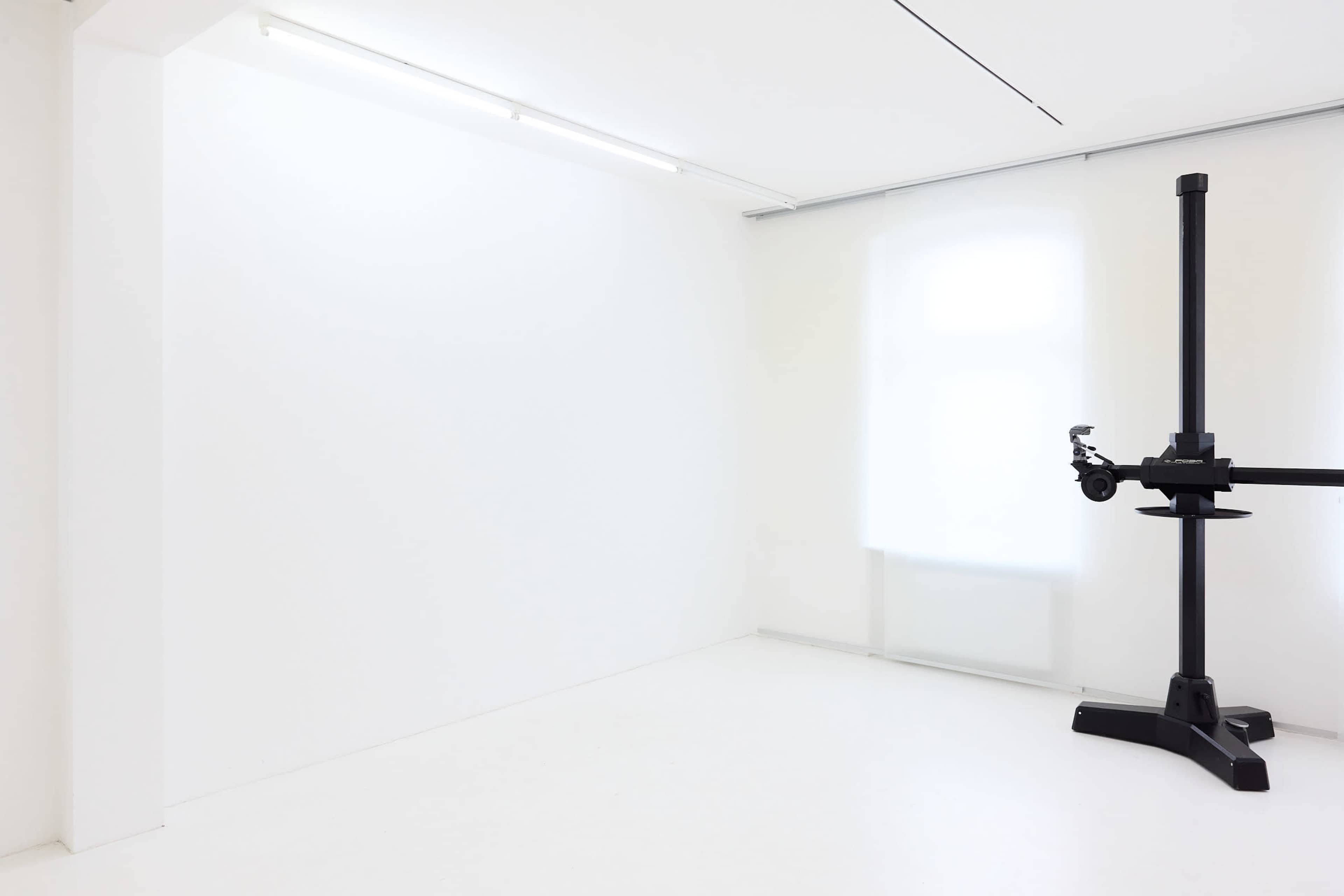 The image shows a minimalist interior with a white wall, a light source from a window, and a black stand or structure in one corner.
