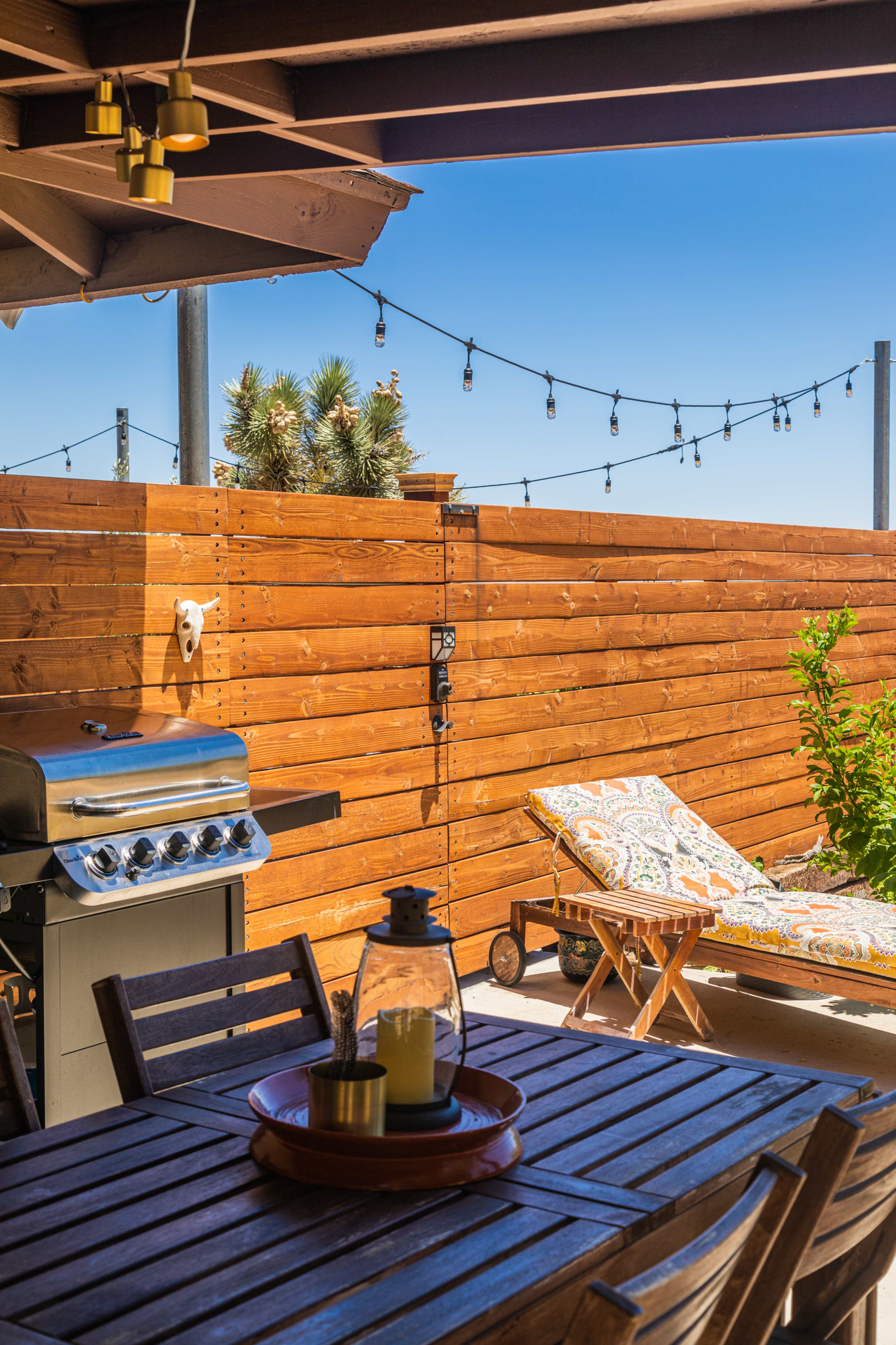 A wooden patio features a grill, dining table, and a lounge chair beneath string lights with a clear blue sky in the background.